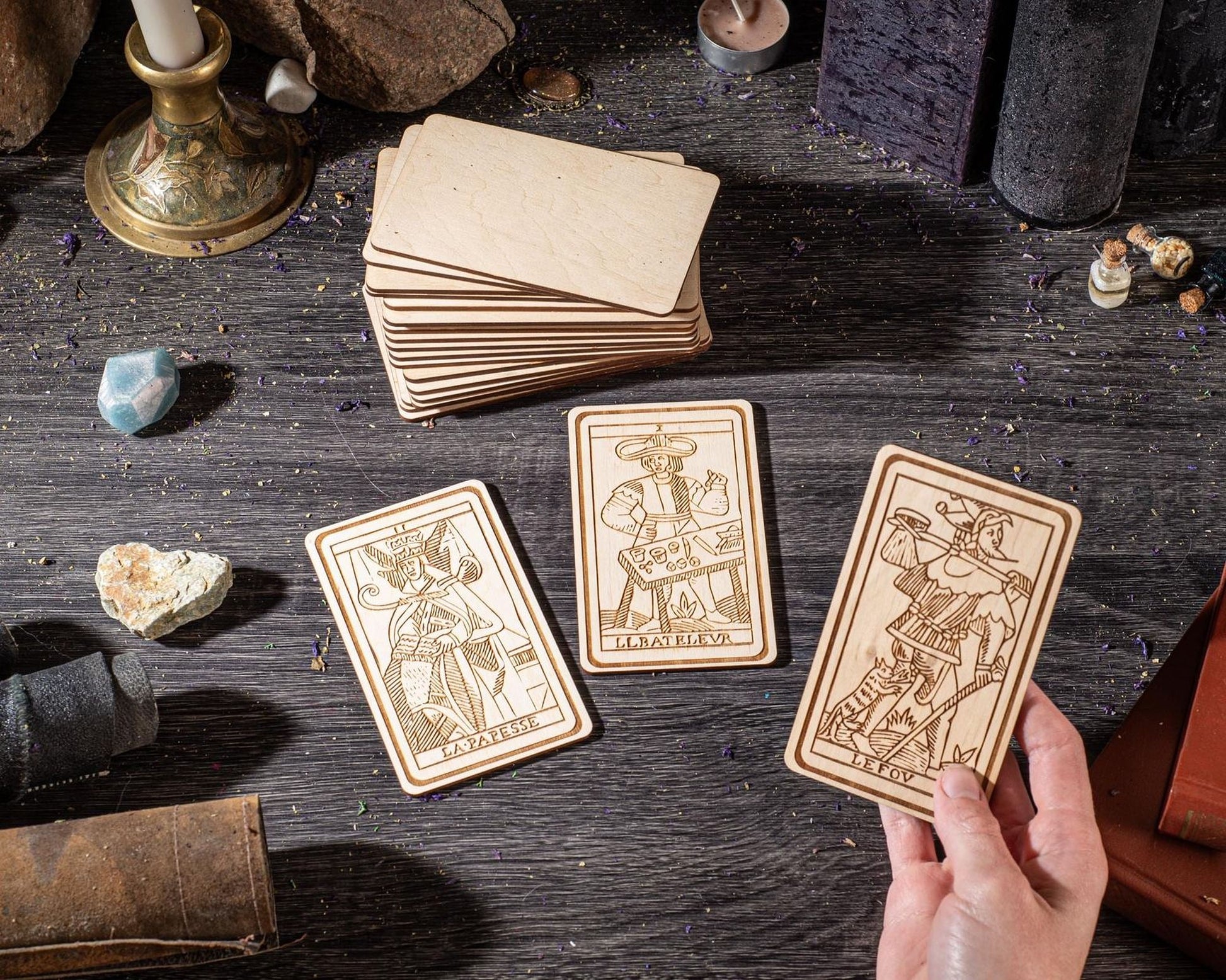 a hand holding a wooden tarot card, surrounded by various objects such as candles, crystals, and other items on a wooden surface.