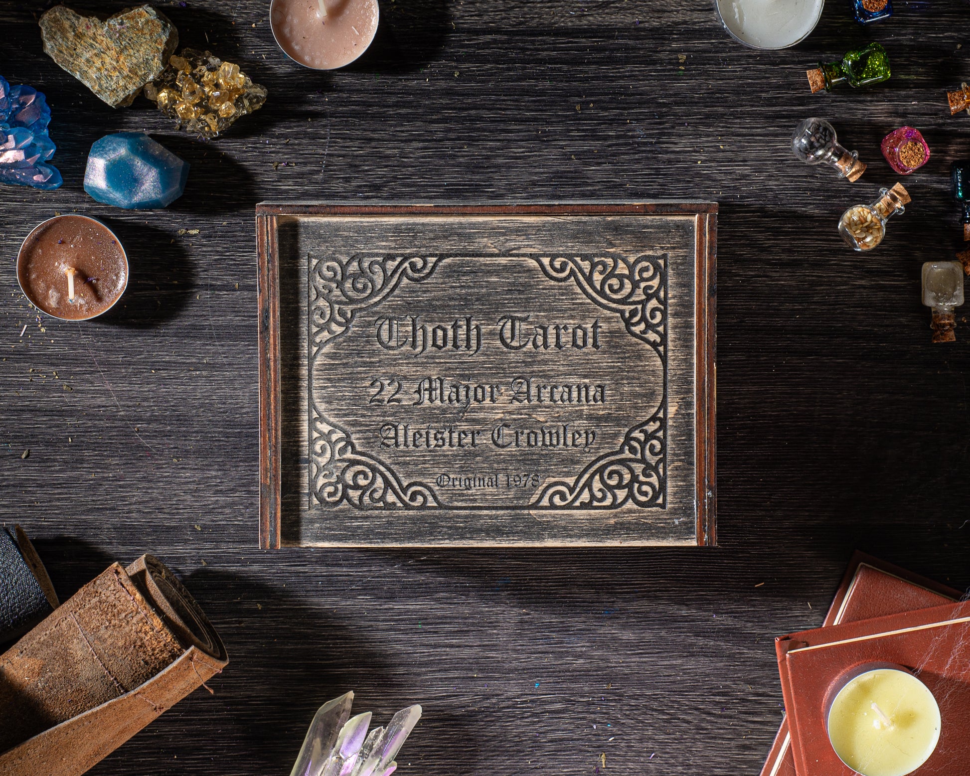 a wooden box with a decorative border, surrounded by various objects such as candles, crystals, and books, all placed on a wooden surface.