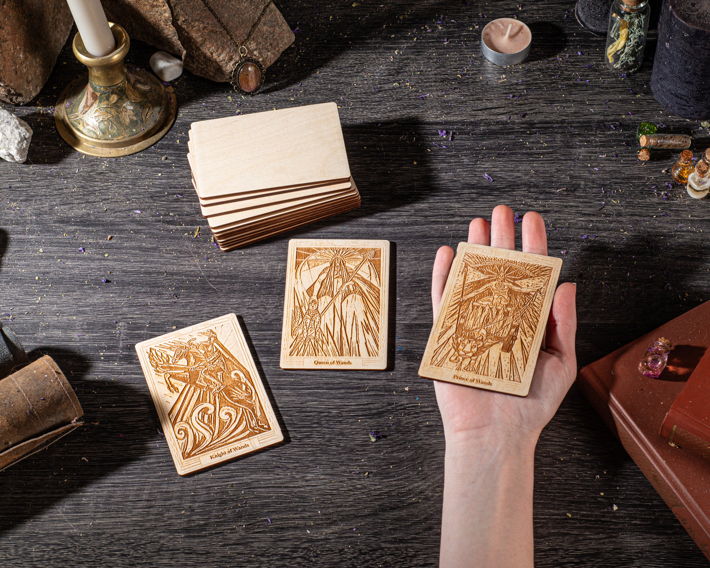 A hand holding three wooden tarot cards with intricate designs, surrounded by various objects such as candles, crystals, and books on a dark wooden surface.
