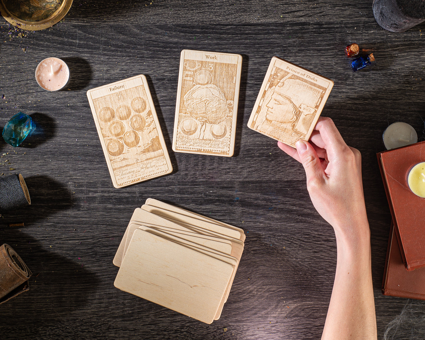 a hand holding a wooden card with a picture of a person on it, surrounded by various objects such as candles, books, and other wooden items on a dark wooden surface.