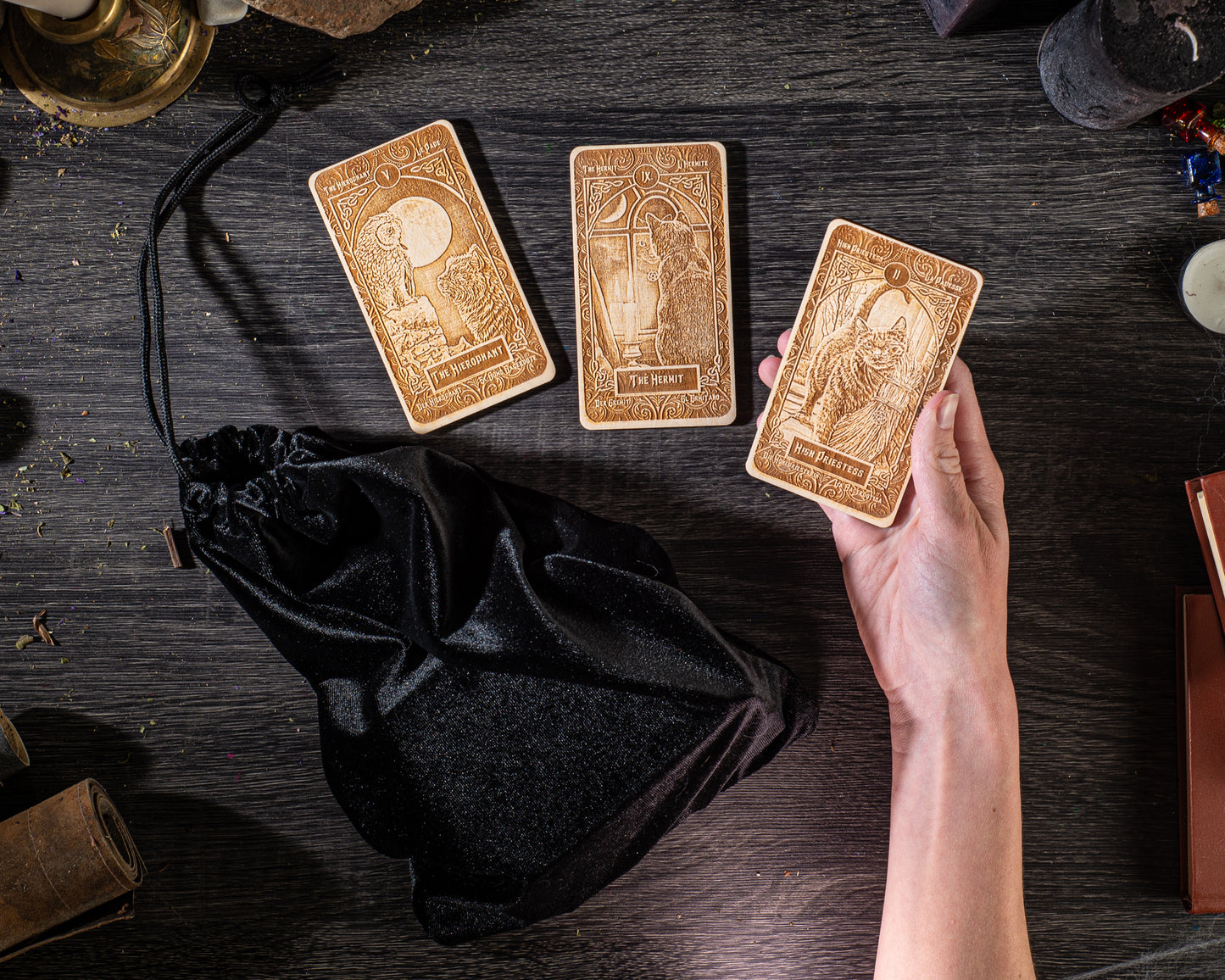 a hand holding three wooden tarot cards on a dark wooden surface, with a black satchel and various objects in the background.