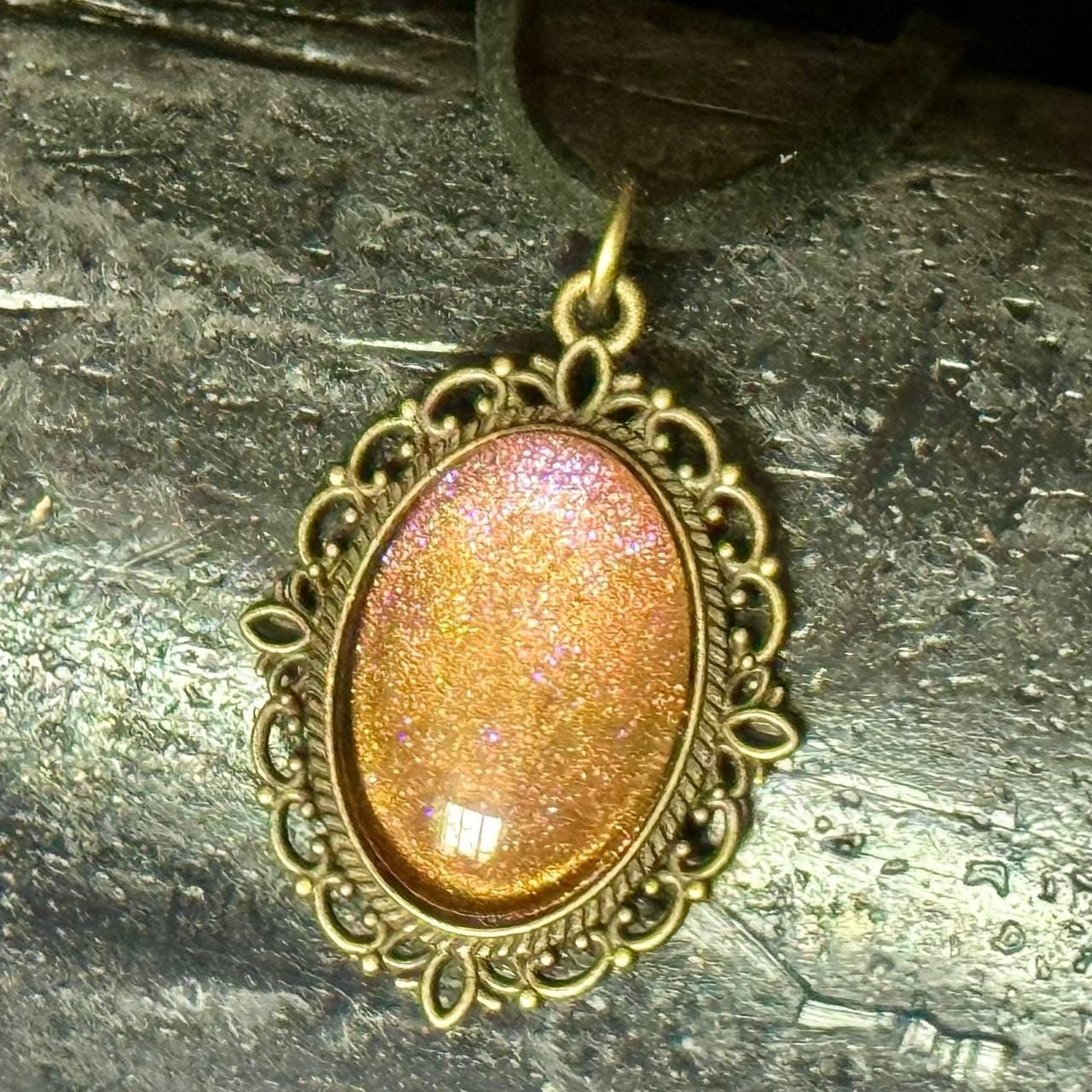 A decorative pendant with a glittery, oval-shaped stone or gemstone in the center, surrounded by an ornate, gold-colored frame.