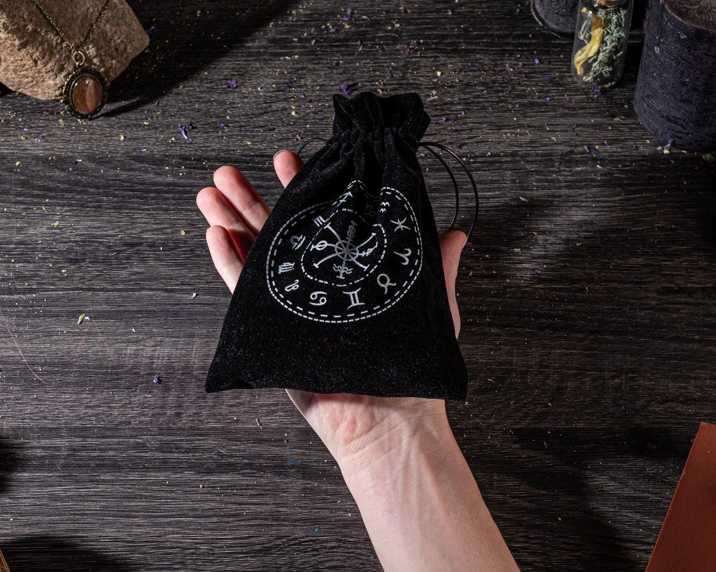 A hand holding a black pouch with a circular design on it, placed on a wooden surface with various objects in the background.