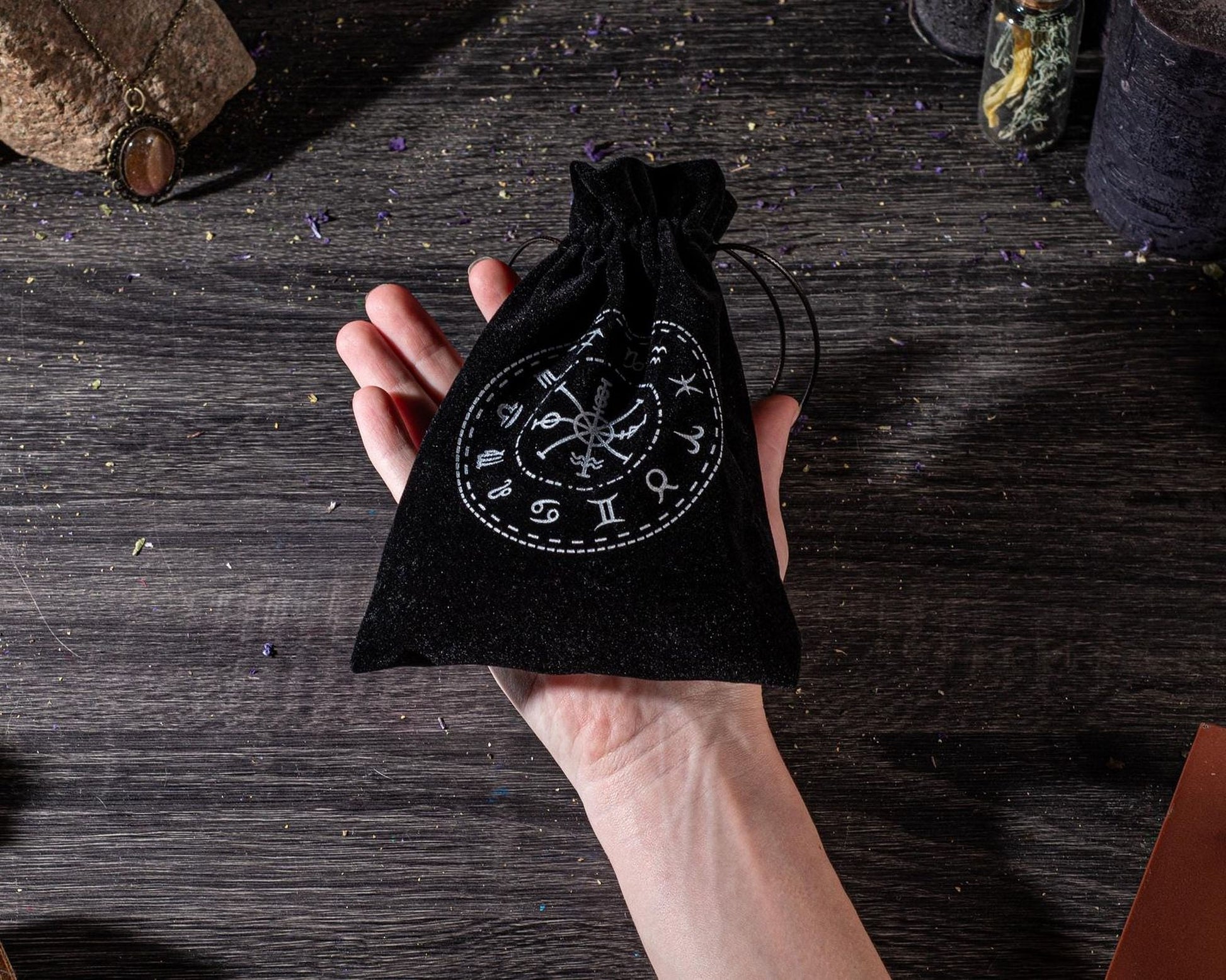 A hand holding a black pouch with a circular design on it, placed on a wooden surface with various objects in the background.