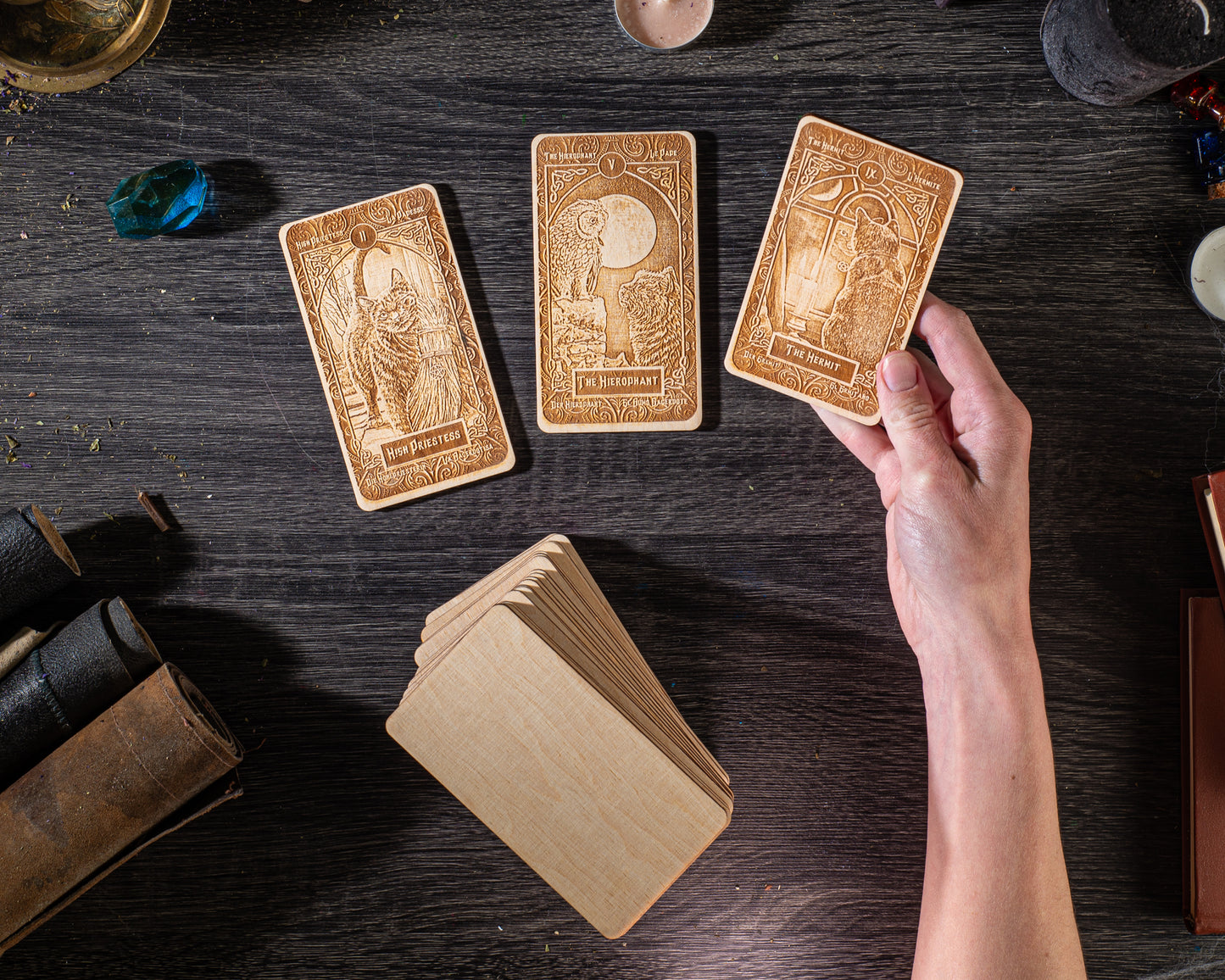 A hand is holding three wooden tarot cards on a dark wooden surface, surrounded by various objects such as books, crystals, and candles.