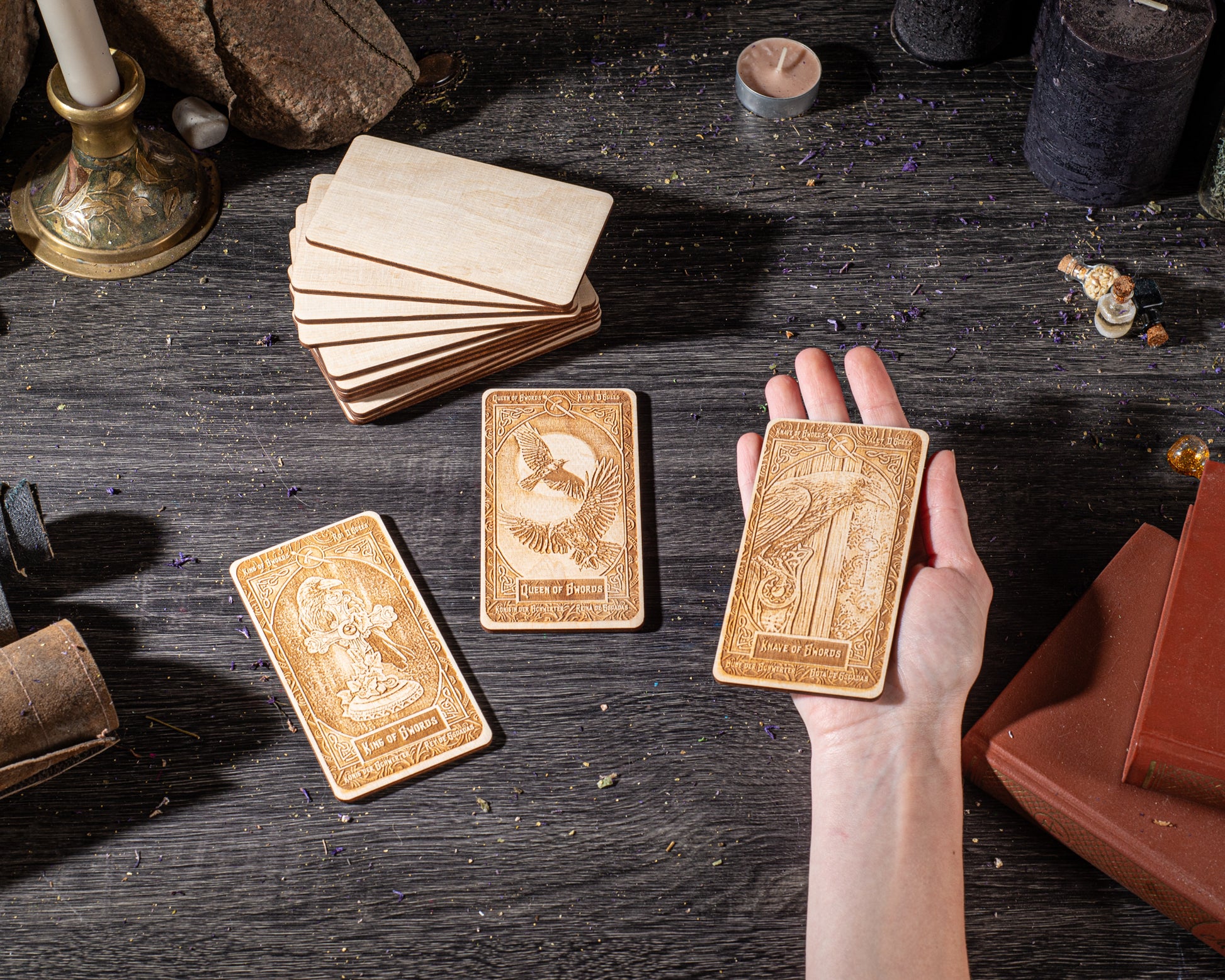 A hand is holding three wooden tarot cards on a dark wooden surface, surrounded by various objects such as candles, books, and other items.