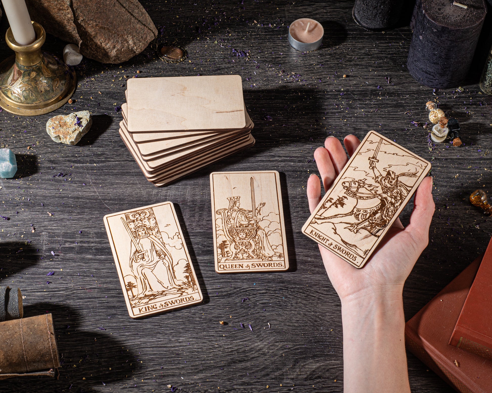 A hand holding three wooden tarot cards with intricate designs, surrounded by various objects such as crystals, candles, and other items on a dark wooden surface.