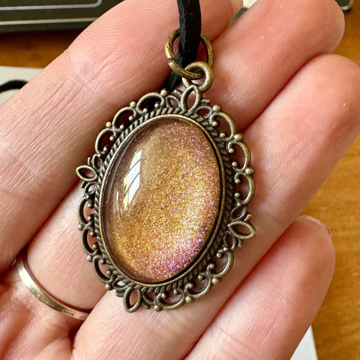 A hand holding a decorative pendant with a glittery stone or gemstone in the center, surrounded by an ornate metal frame.