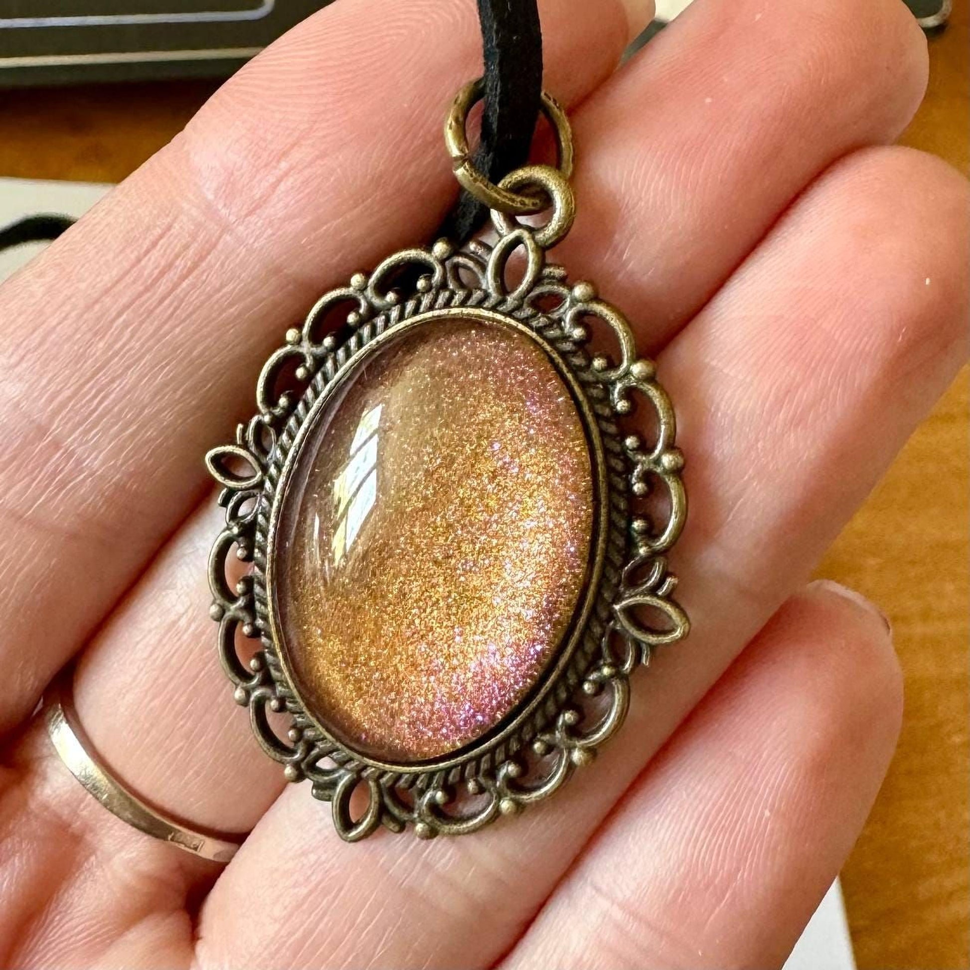 A hand holding a decorative pendant with a glittery stone or gemstone in the center, surrounded by an ornate metal frame.