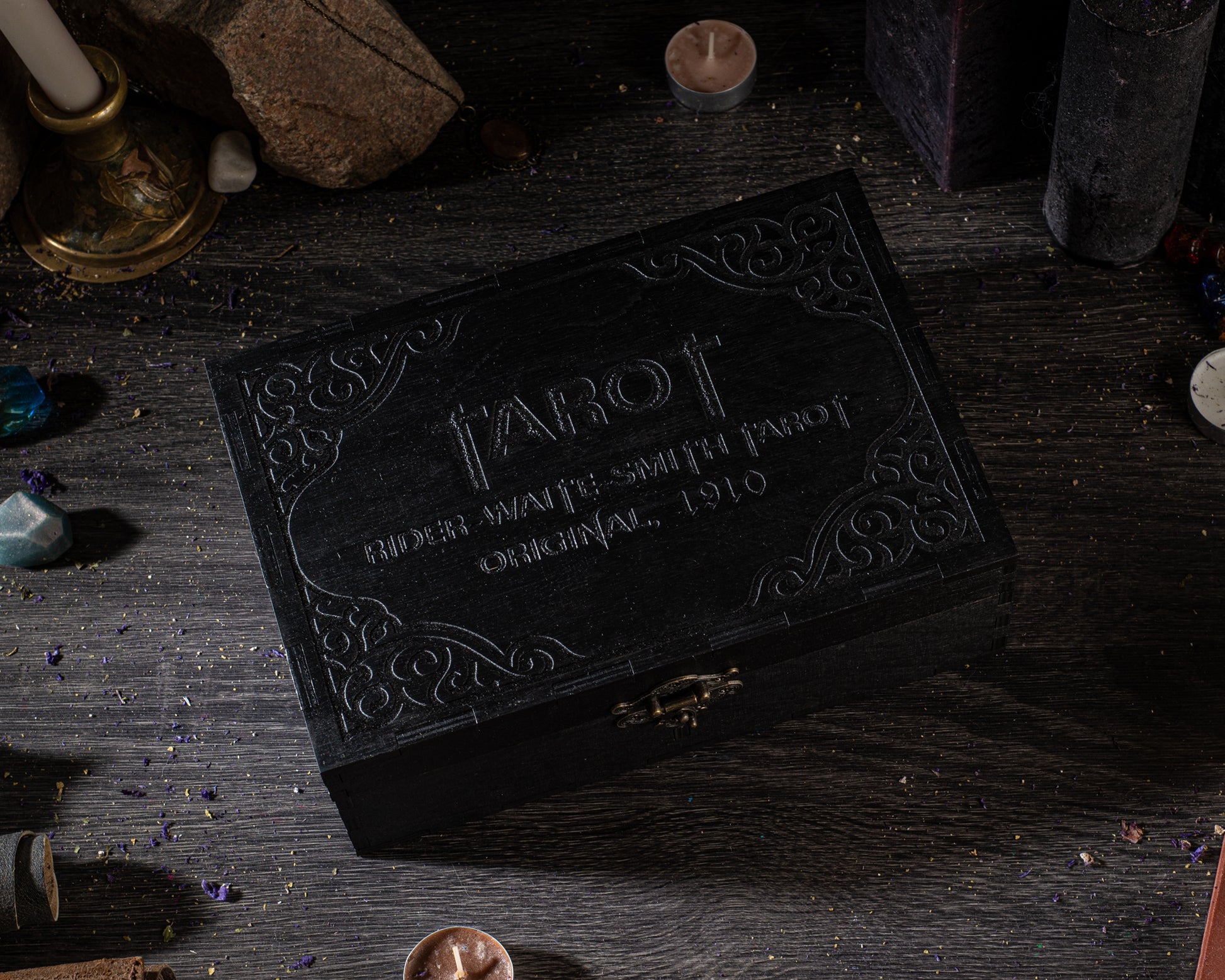 A black, ornately decorated box with the word "TAROT" embossed on it, sitting on a wooden surface surrounded by various objects such as candles, gemstones, and other items.