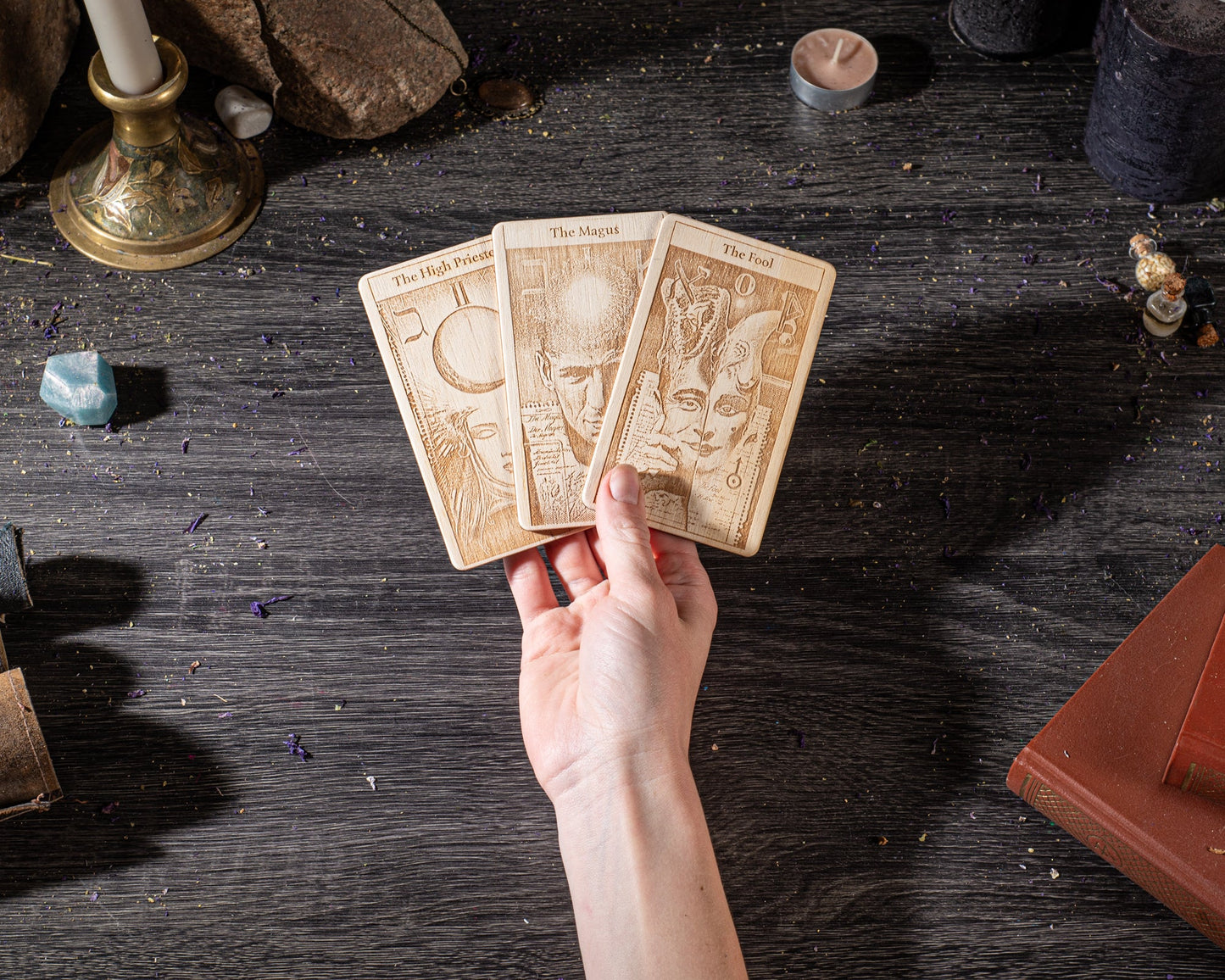 A hand is holding three tarot cards on a dark wooden surface, surrounded by various objects such as candles, crystals, and books.