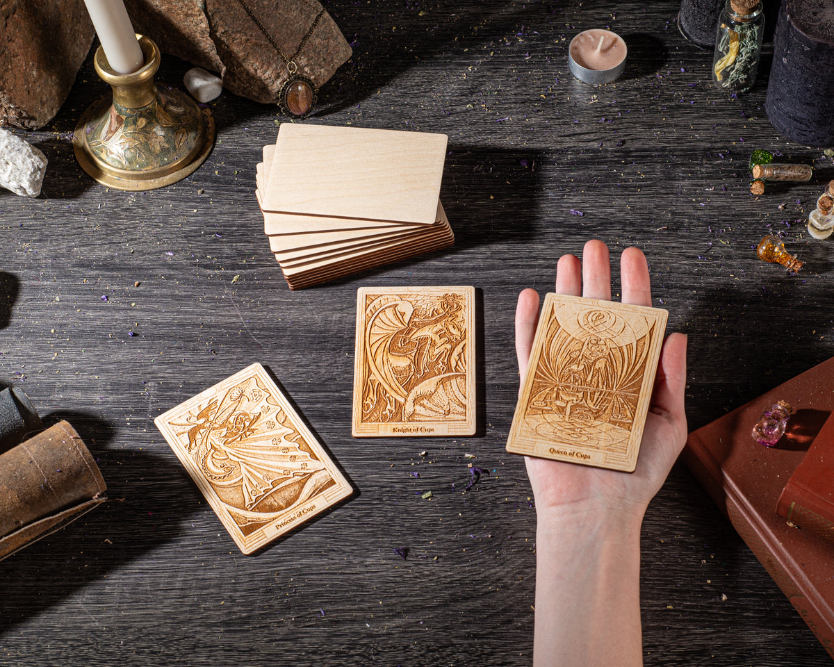 A hand holding three wooden tarot cards with intricate designs, surrounded by various objects such as candles, crystals, and books on a dark wooden surface.