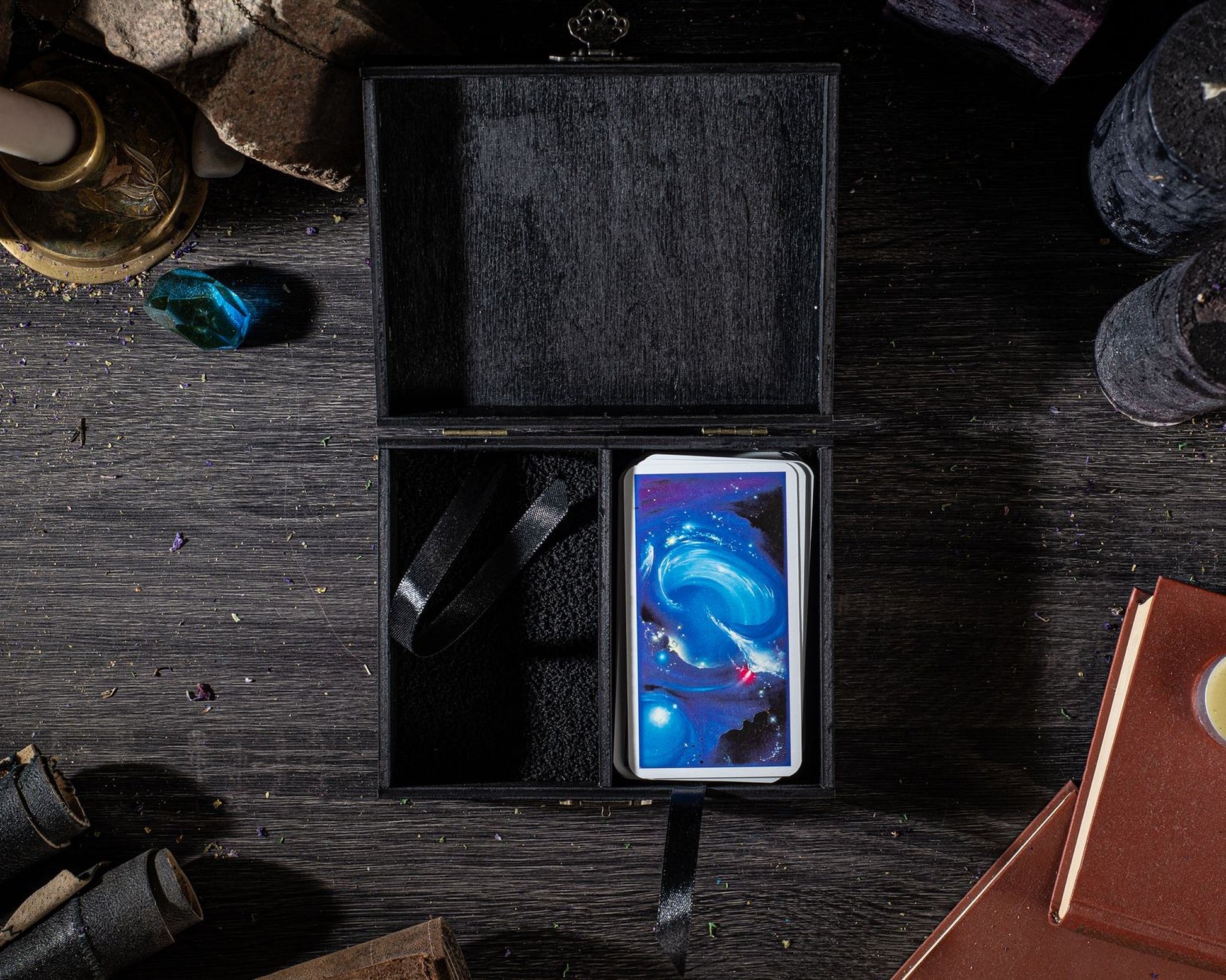 a black wooden box with a colorful, abstract design on its front, placed on a dark wooden surface. Surrounding the box are various objects, including a candle, a book, and a pair of glasses, creating a visually interesting and diverse composition.