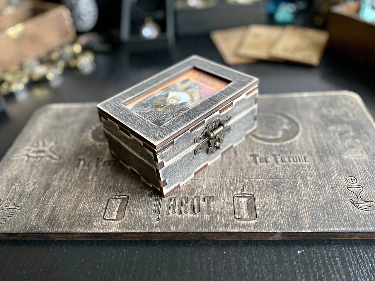 A small wooden box with a picture on it, placed on a wooden surface with a tarot card reading mat underneath.