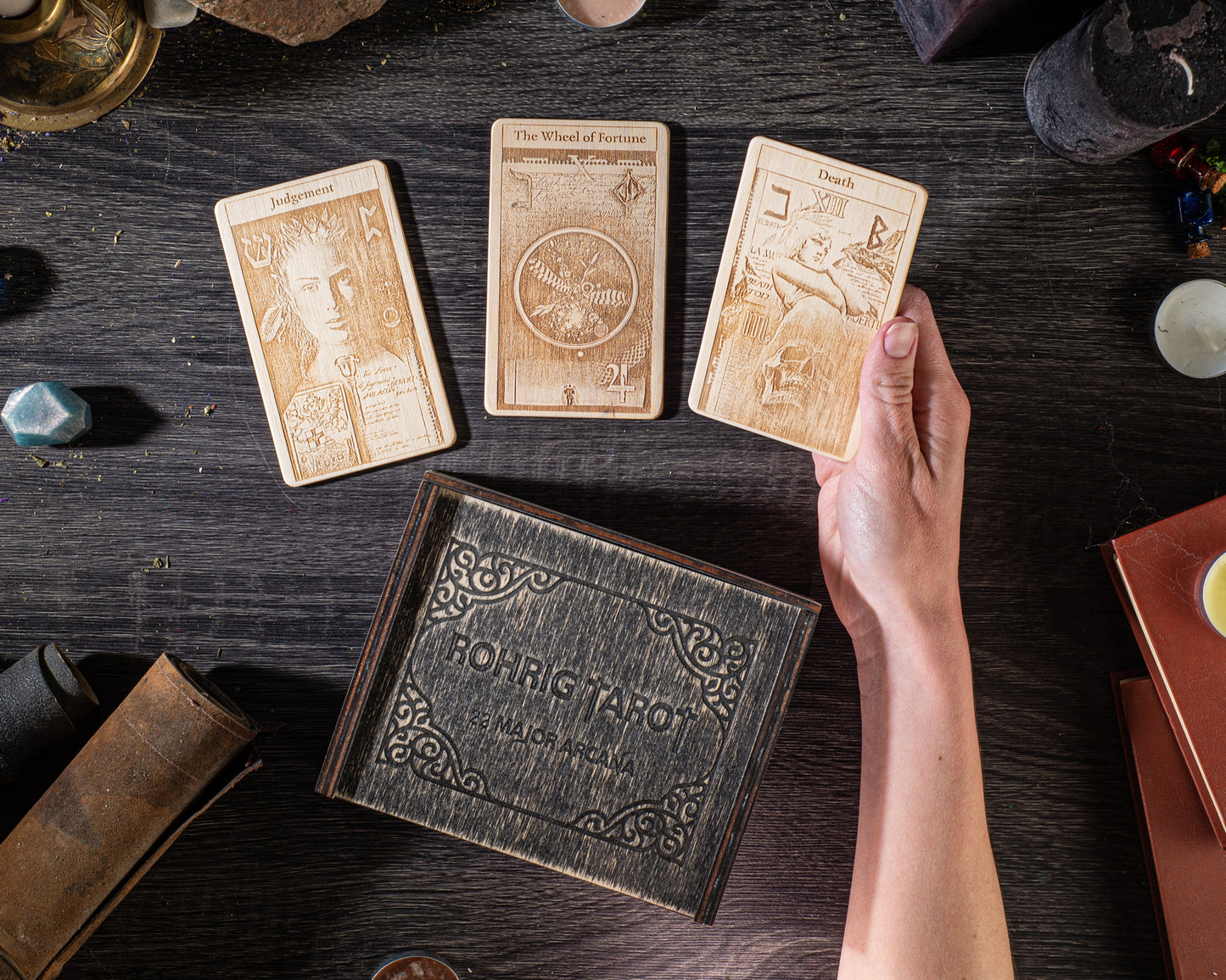a wooden box with ornate designs, a hand holding a wooden card, and various other objects on a dark wooden surface.