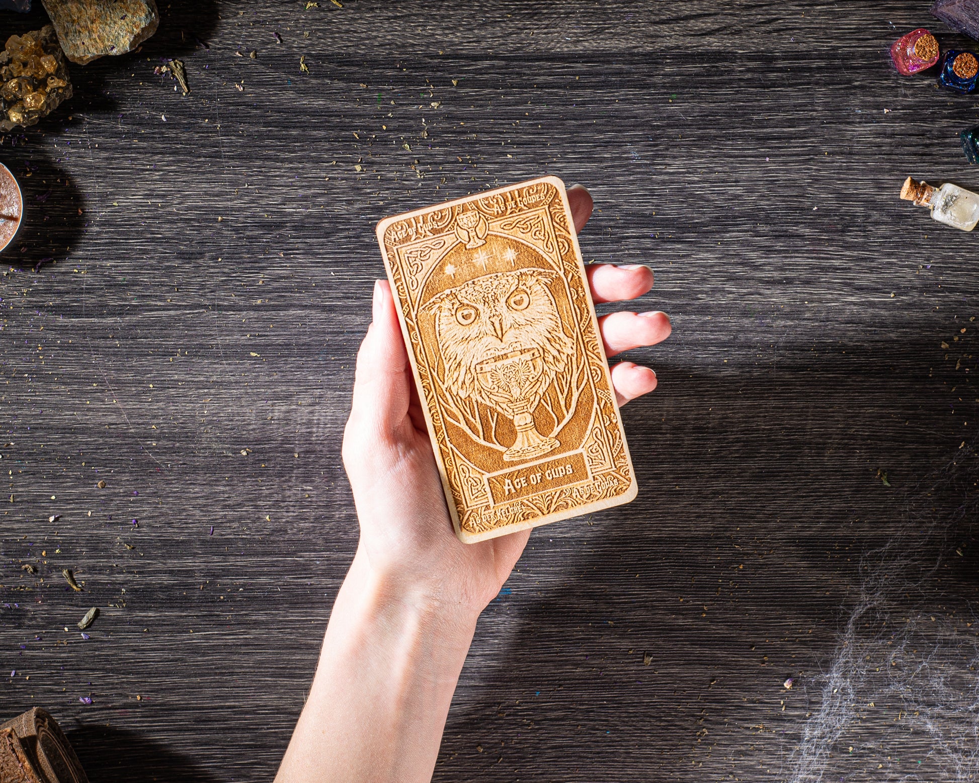 A hand holding a wooden object with an intricate design, possibly a tarot card, against a dark wooden background.