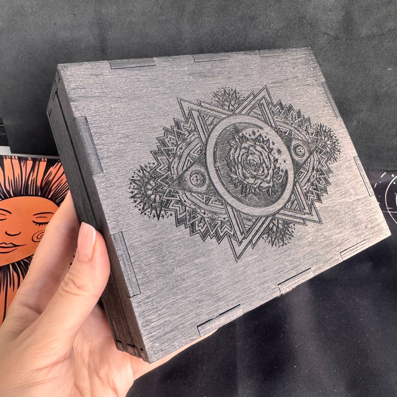 A hand is holding a wooden box with an intricate design featuring a crescent moon and a rose.