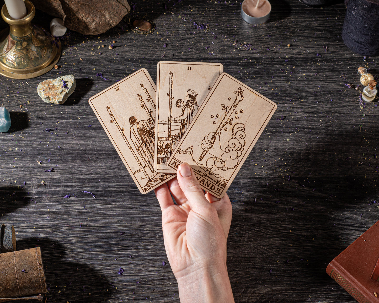 A hand is holding three wooden tarot cards against a dark background, with various objects such as crystals, candles, and books scattered around.
