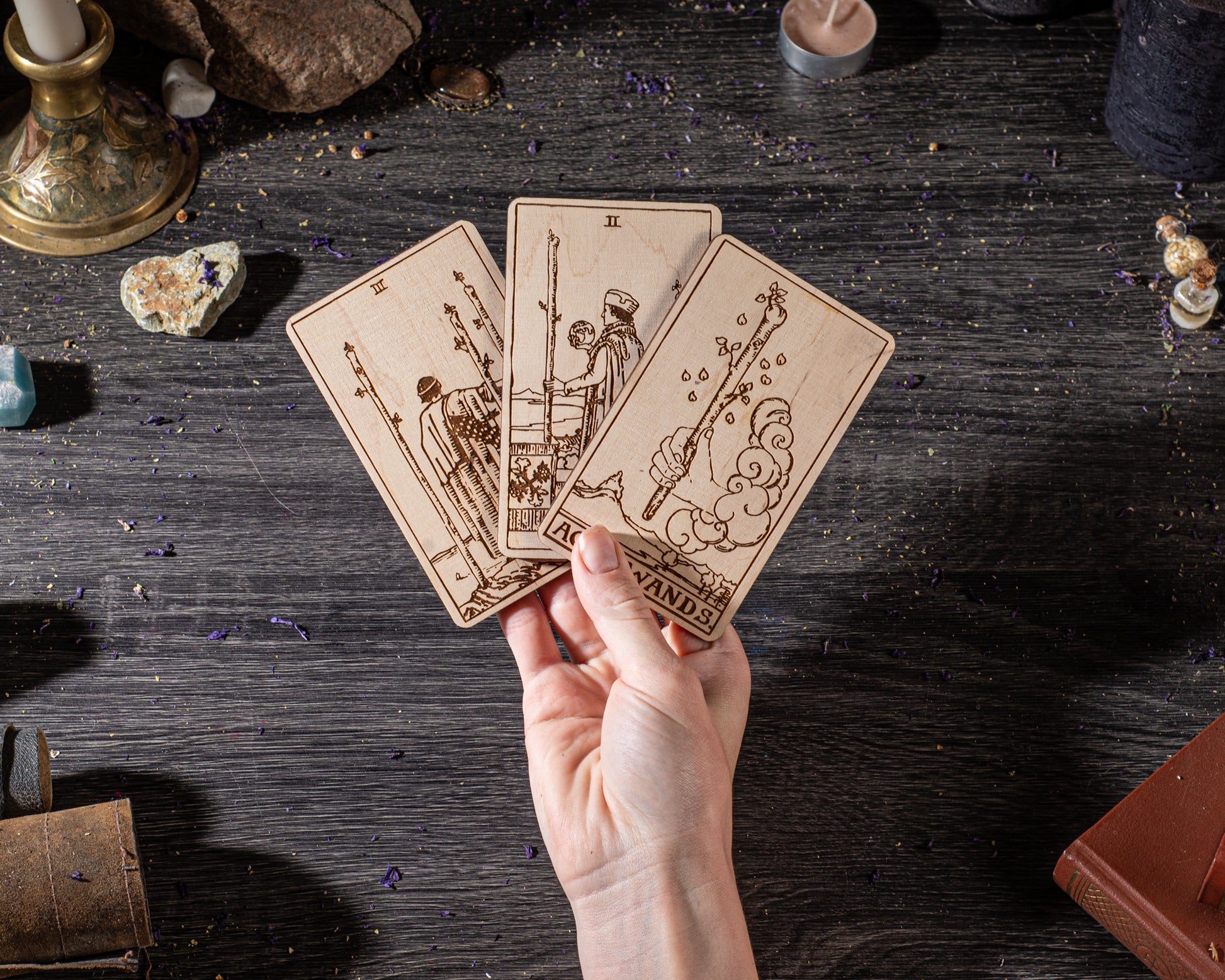 A hand is holding three wooden tarot cards against a dark background, with various objects such as crystals, candles, and books scattered around.