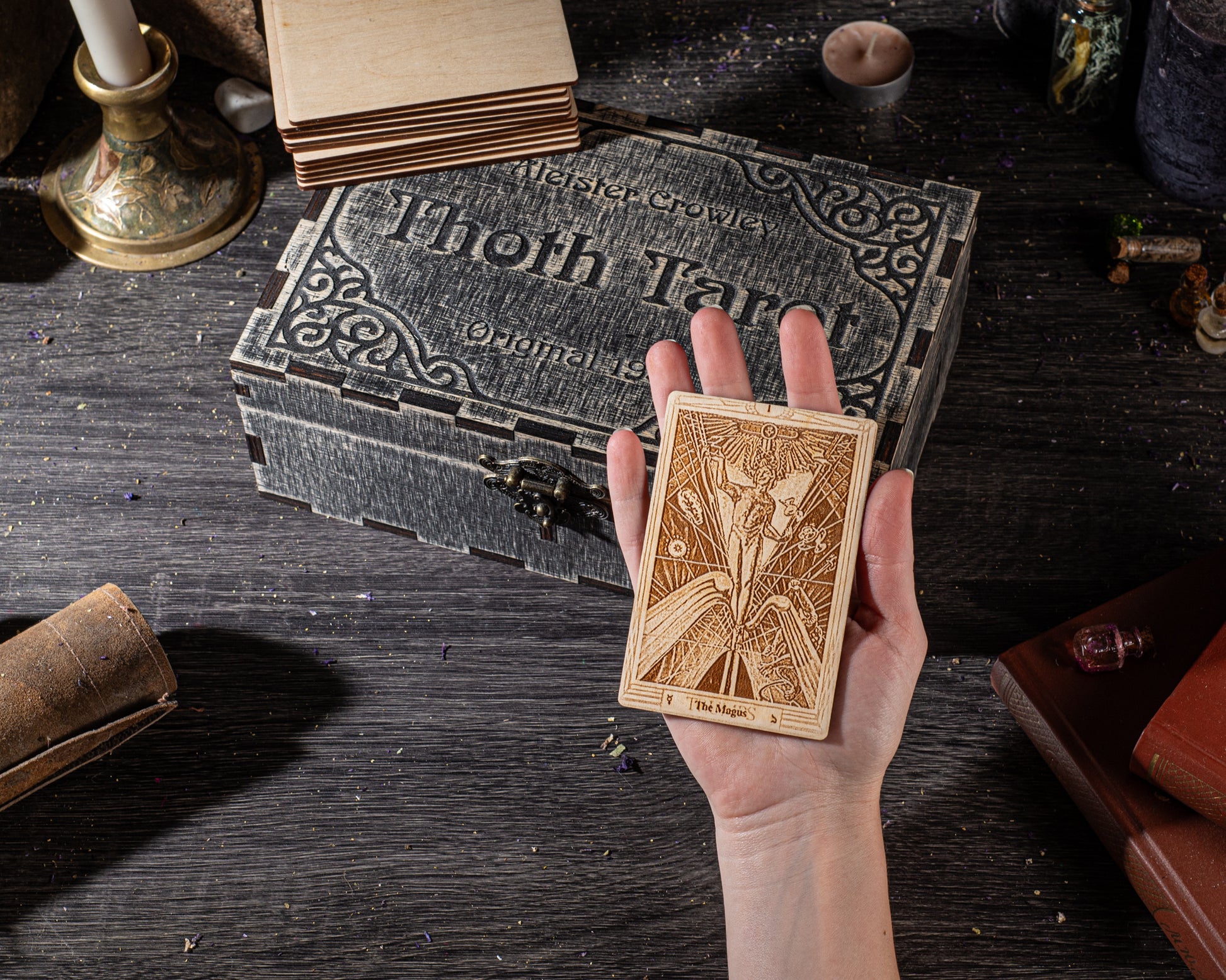 A hand holding a wooden tarot card in front of a stack of books and a wooden box with a decorative design.