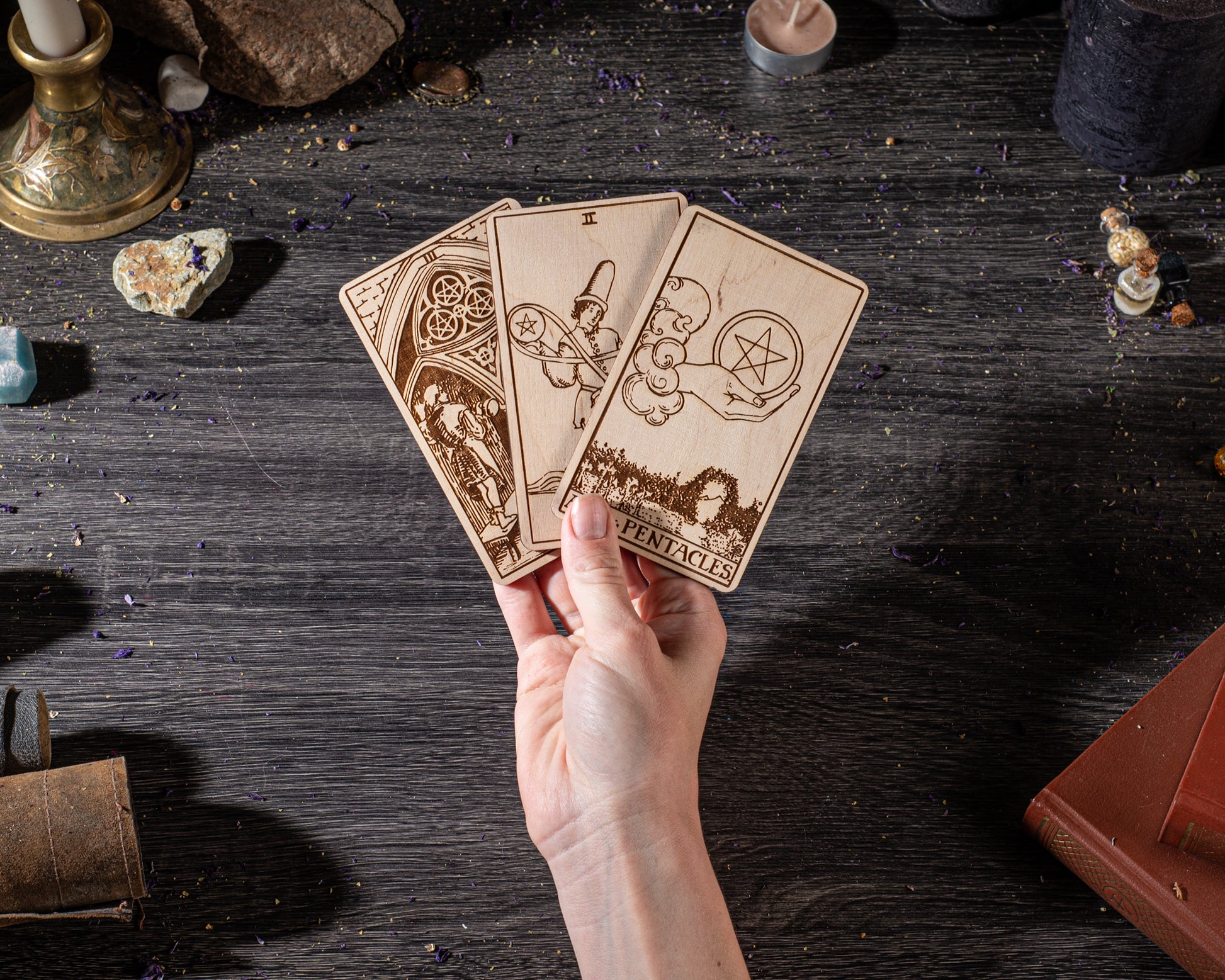 A hand is holding three wooden tarot cards against a dark background, surrounded by various objects such as crystals, candles, and books.