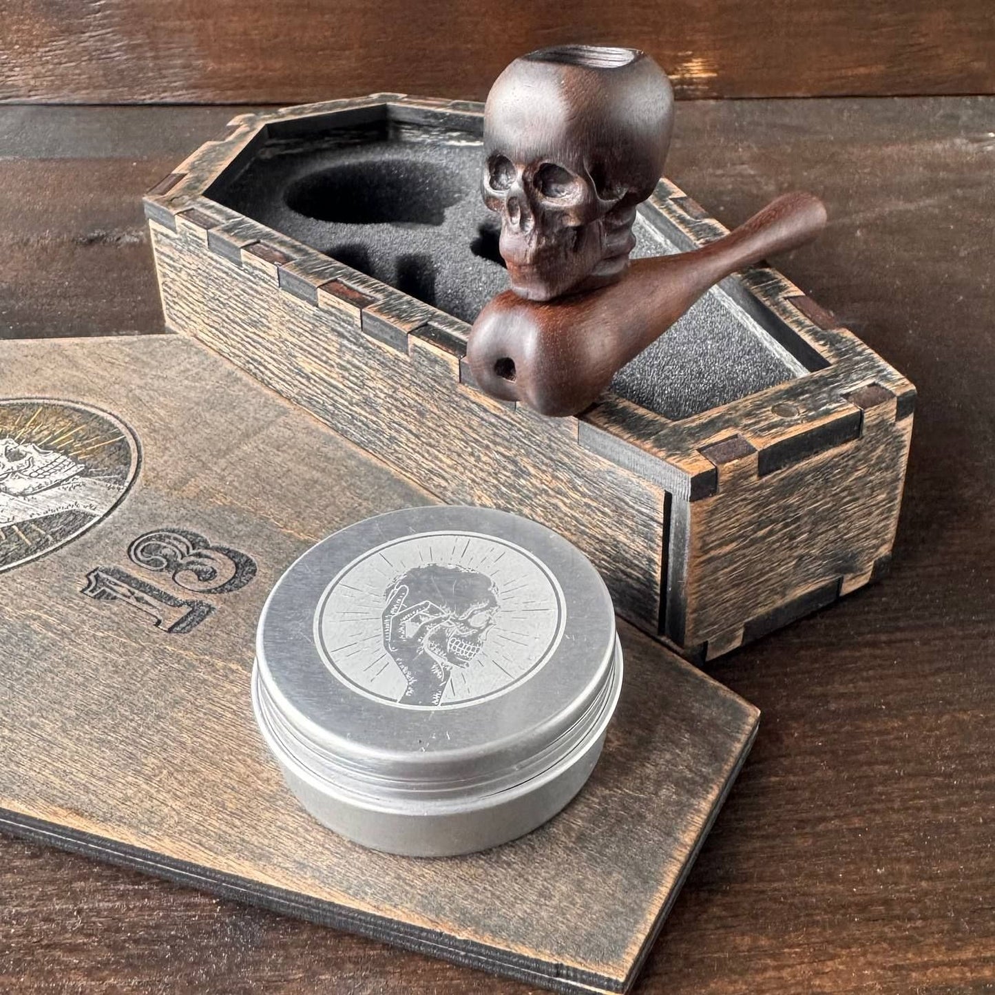 a wooden pipe with a skull design, a metal tin, and a wooden box with a skull design, all placed on a wooden surface.