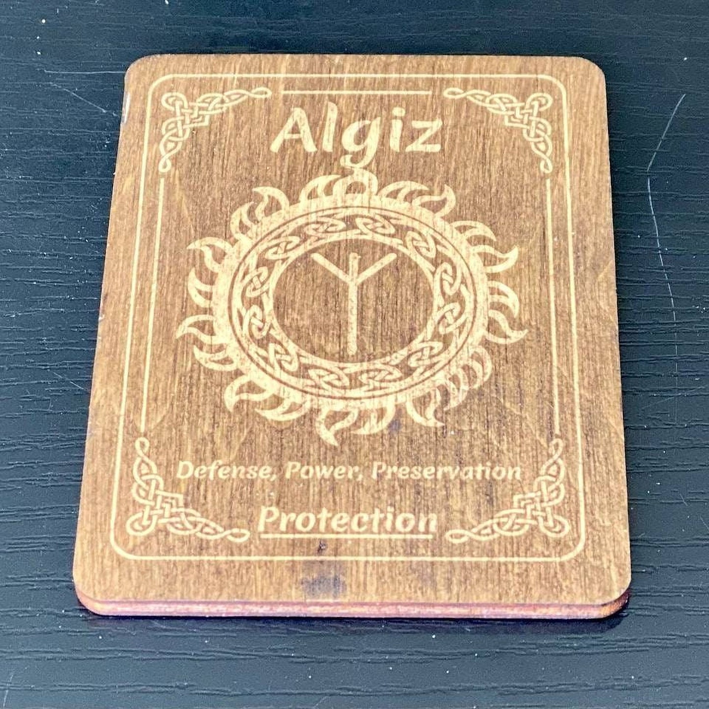 A wooden coaster with the name "Algiz" and a design featuring a sun or flame motif, along with the text "Defense, Power, Preservation" and "Protection".