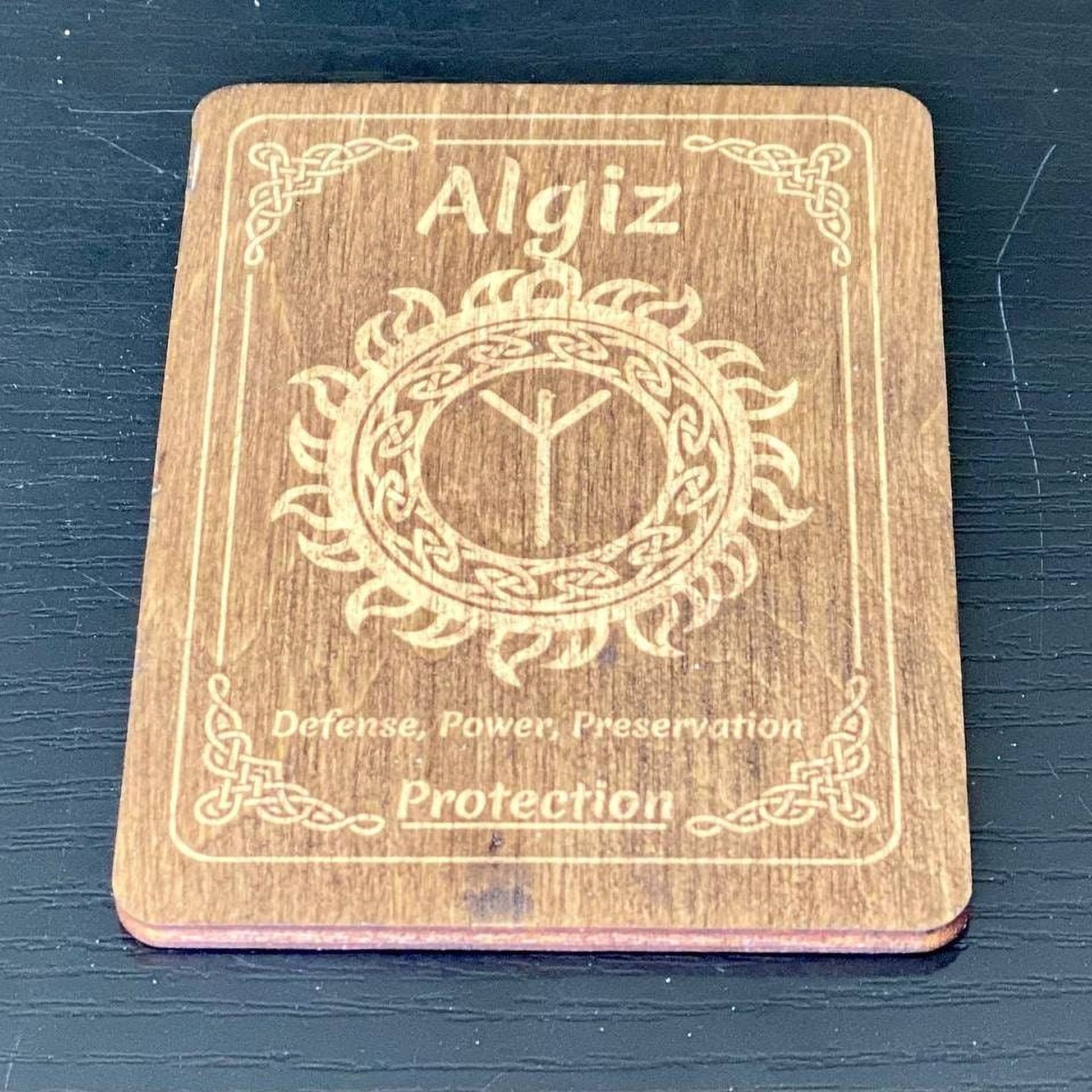 A wooden coaster with the name "Algiz" and a design featuring a sun or flame motif, along with the text "Defense, Power, Preservation" and "Protection".