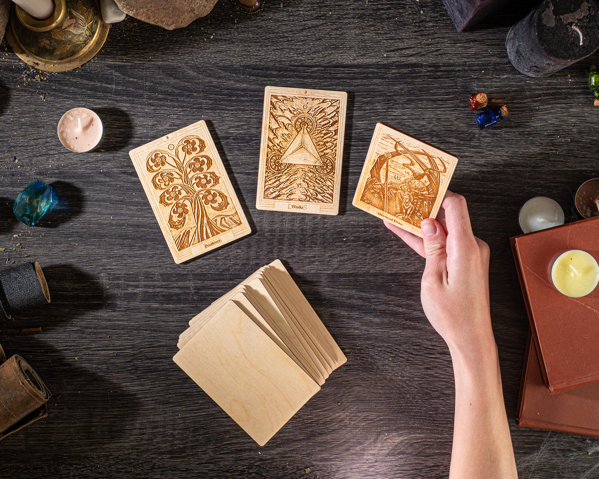 a hand holding a wooden card with an intricate design, surrounded by other wooden cards and objects on a dark wooden surface.