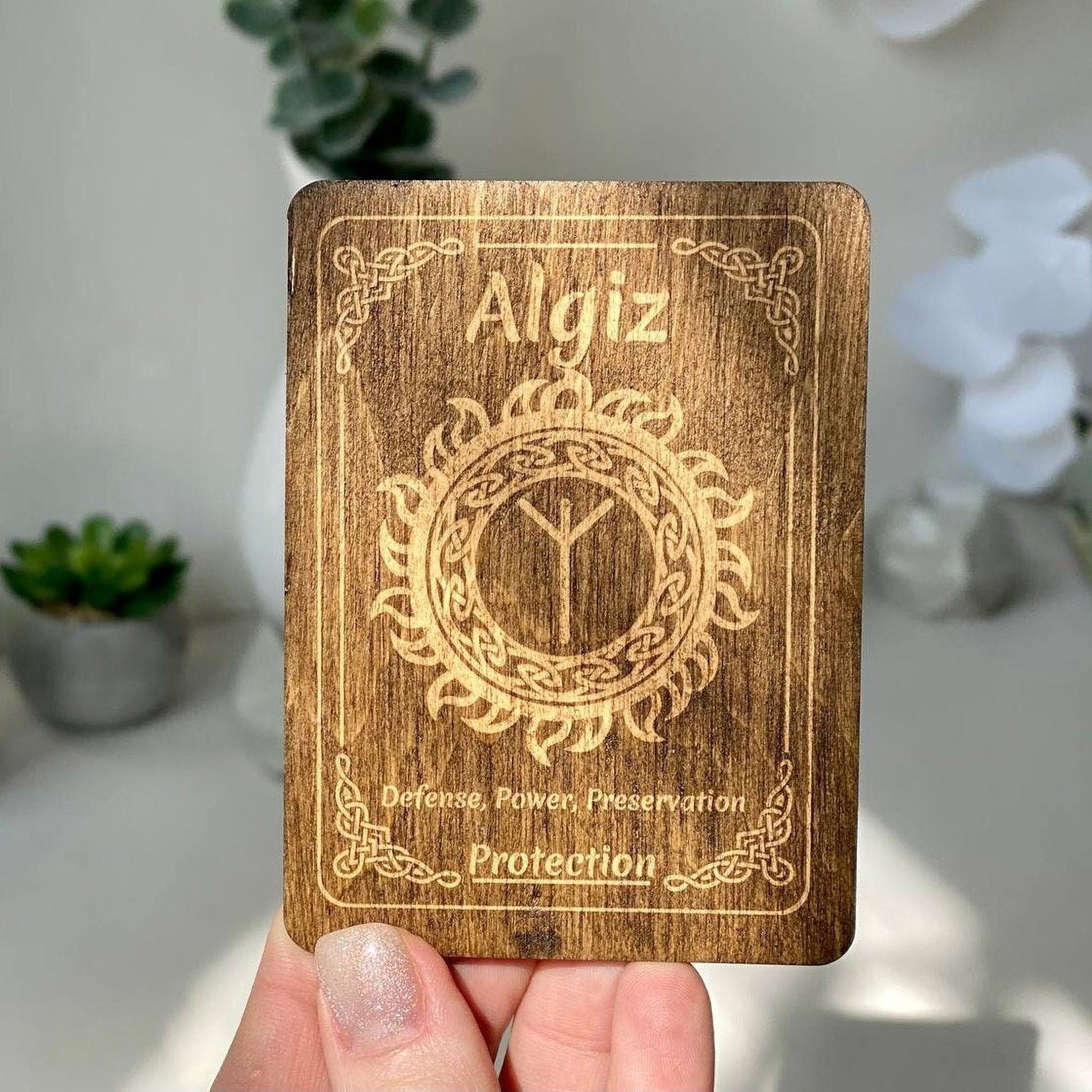 A wooden card with the name Algiz and a design on it is being held up against a blurred background.