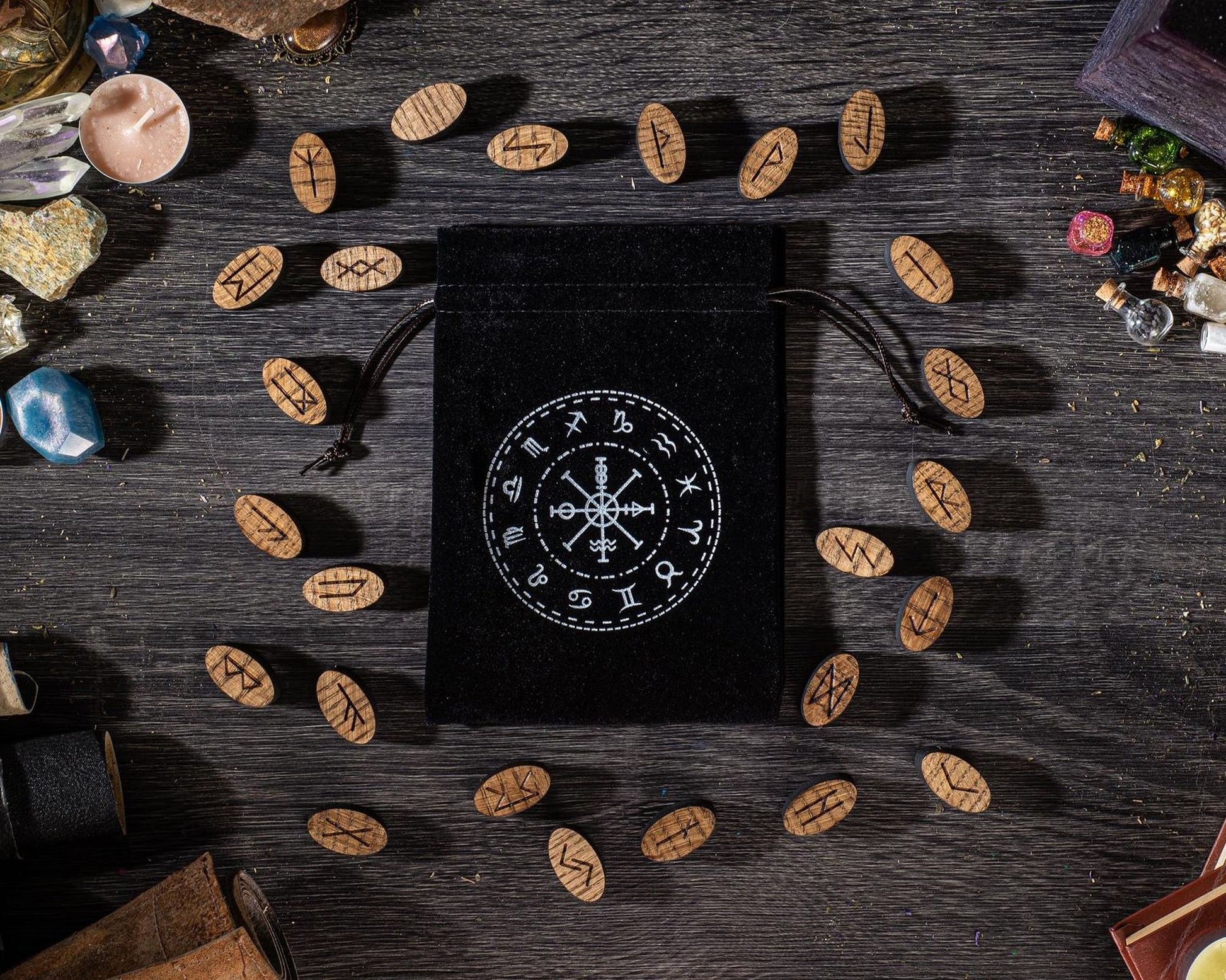 a black pouch with a white design on it, surrounded by various objects such as crystals, stones, and wooden beads on a wooden surface.