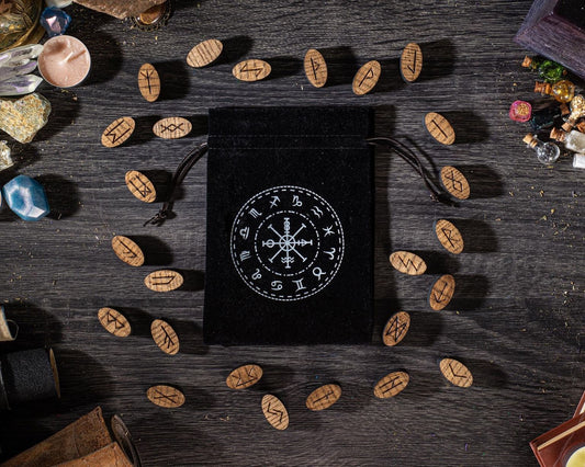 a black pouch with a white design on it, surrounded by various objects such as crystals, stones, and wooden beads on a wooden surface.