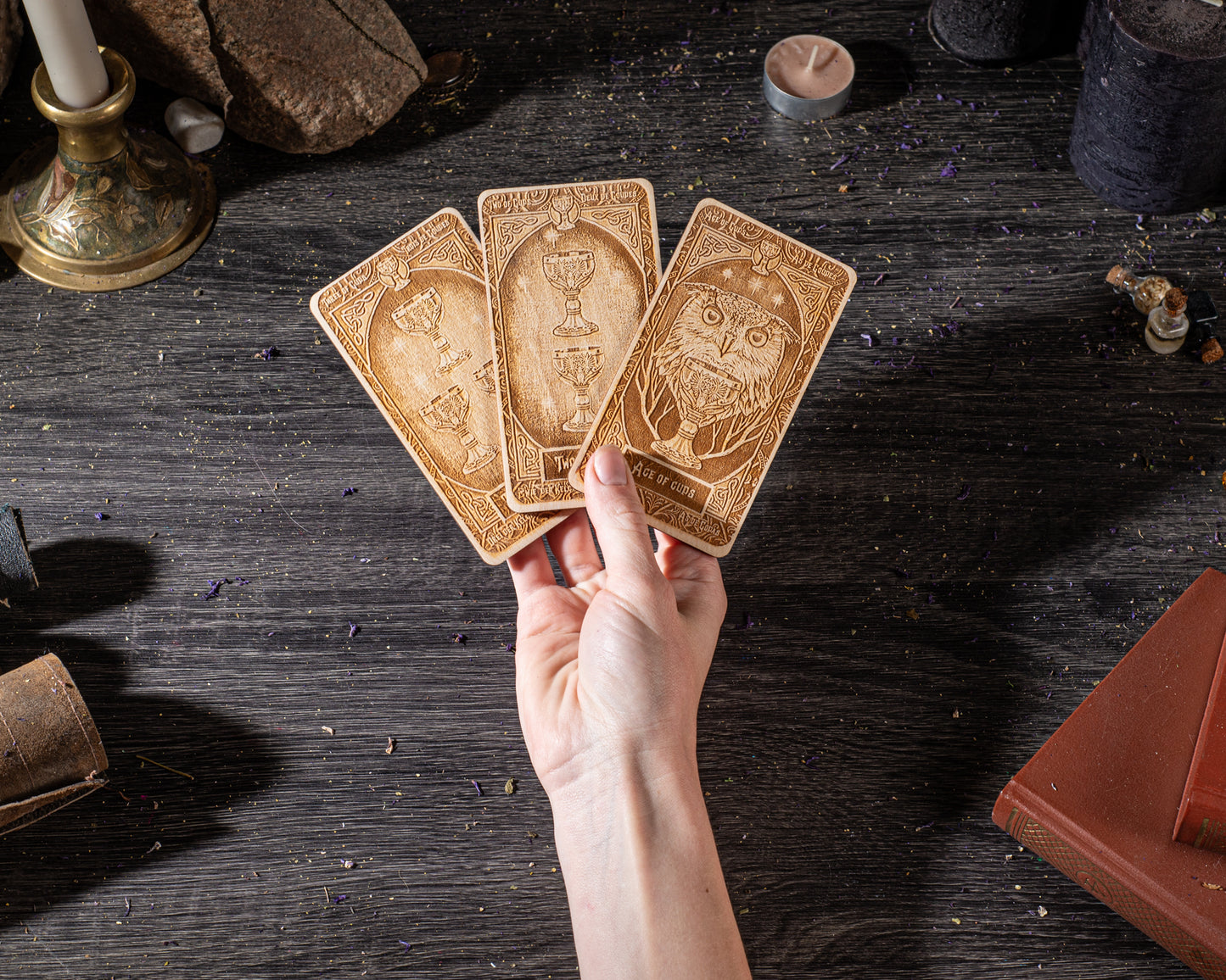 A hand is holding three tarot cards on a dark wooden surface, surrounded by various objects such as candles, books, and crystals.
