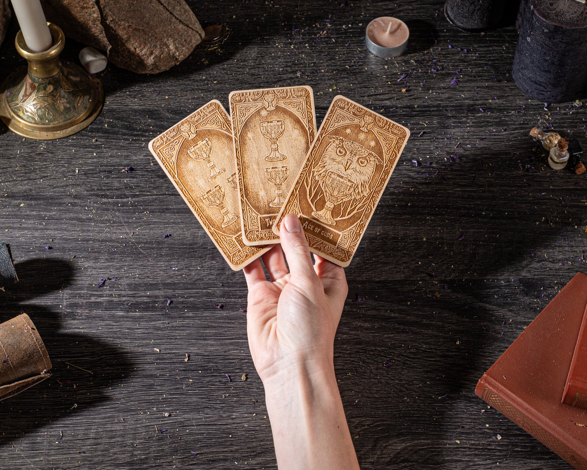 A hand is holding three tarot cards on a dark wooden surface, surrounded by various objects such as candles, books, and crystals.