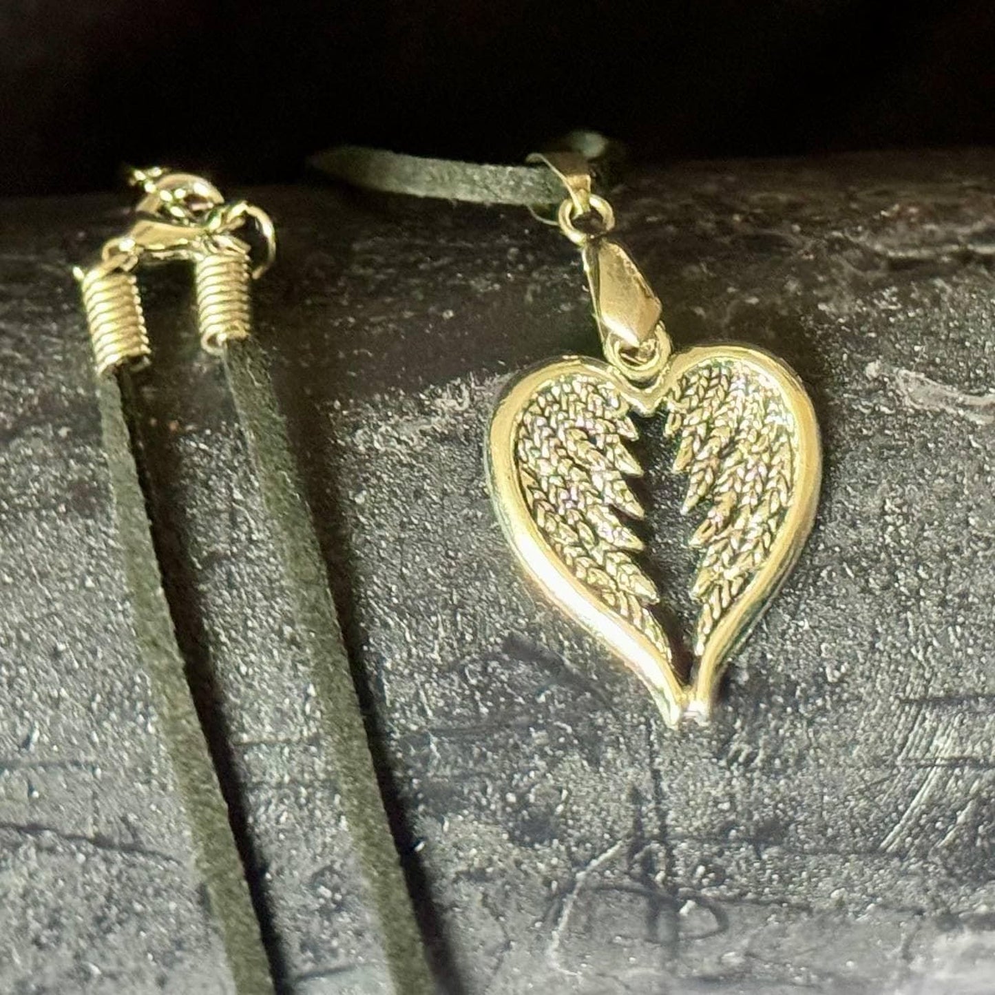 A heart-shaped pendant with a metallic, winged design is displayed on a dark surface, accompanied by a black leather cord.