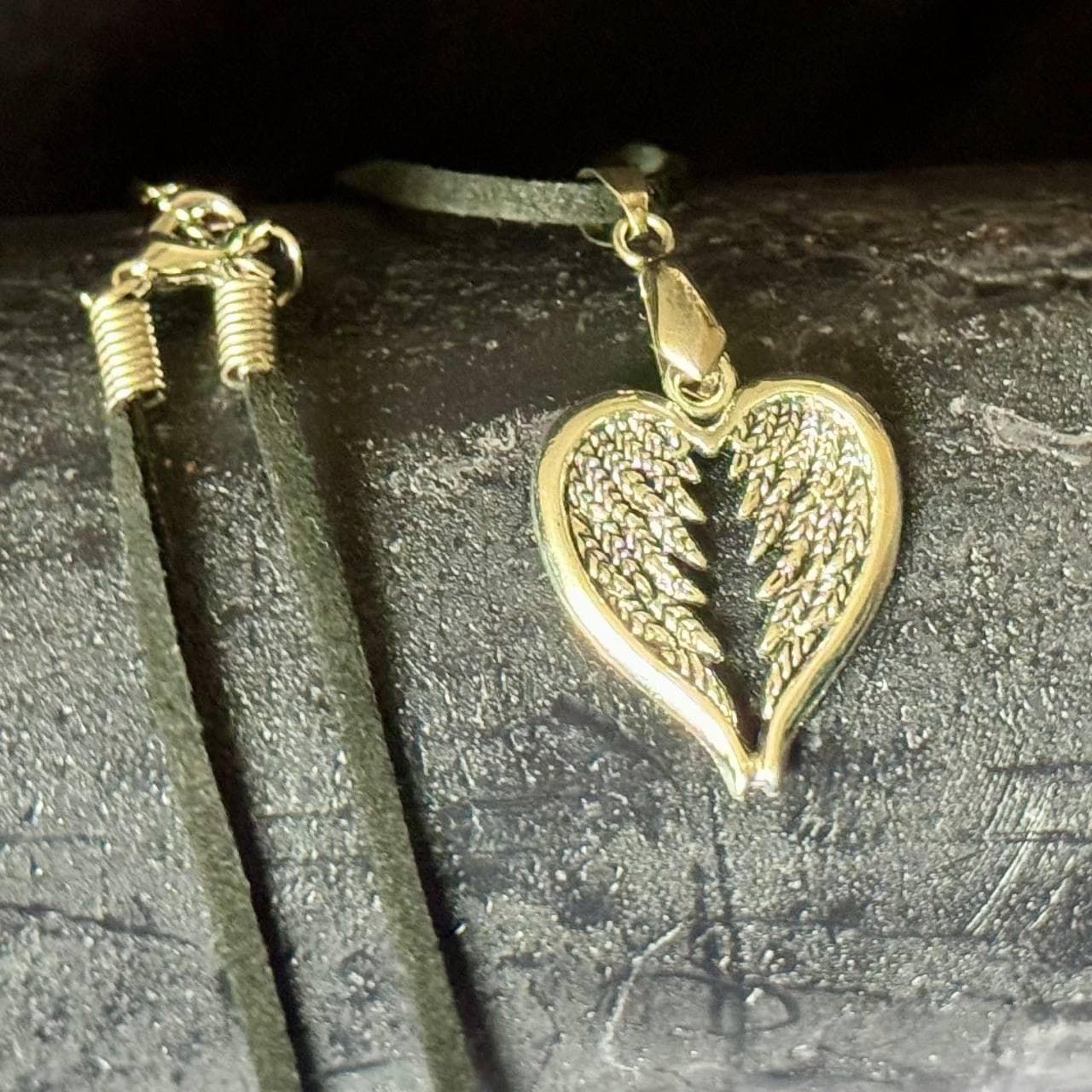 A heart-shaped pendant with a metallic, winged design is displayed on a dark surface, accompanied by a black leather cord.
