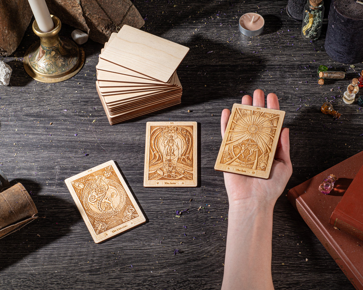 A hand is holding three wooden tarot cards against a dark wooden background, surrounded by various objects such as candles, books, and crystals.