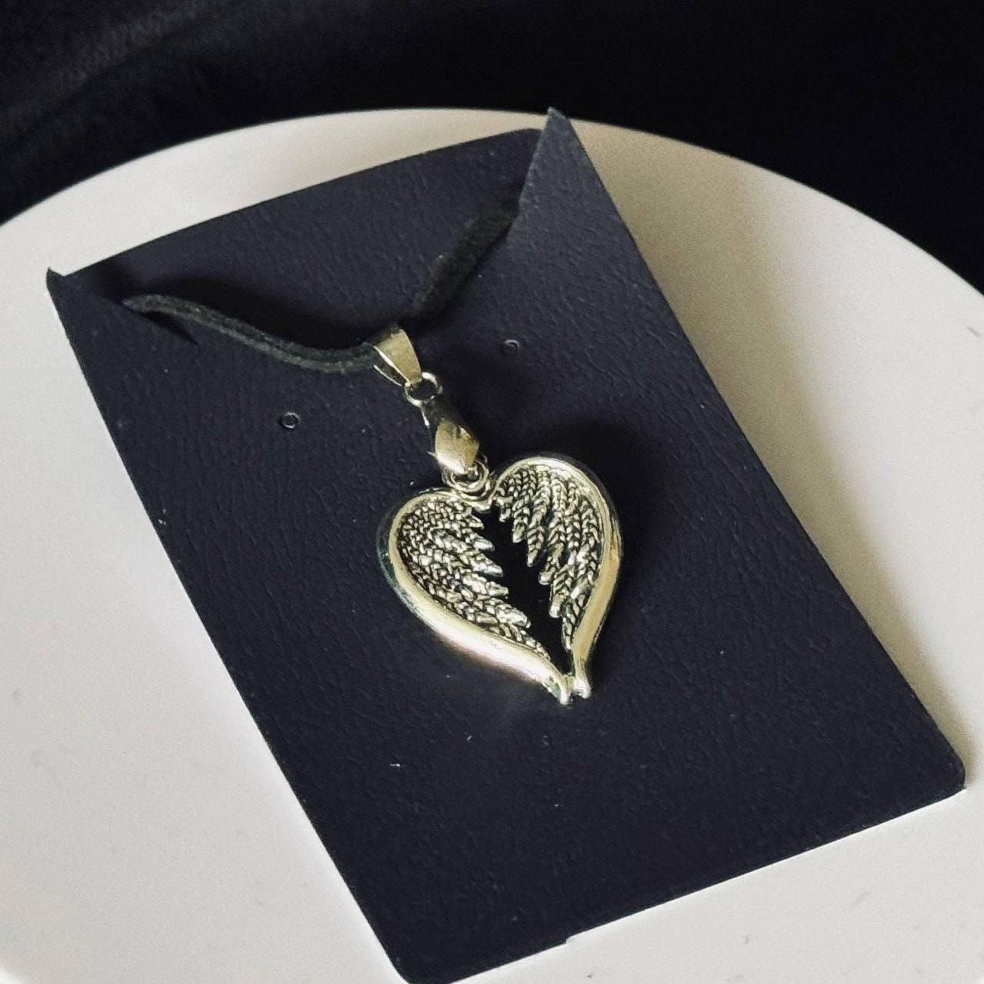 A silver heart-shaped pendant with wings is displayed on a black card.