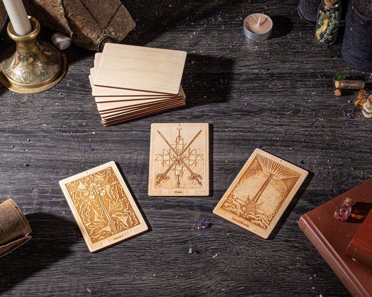 three wooden tarot cards with intricate designs, placed on a dark wooden surface. There are also various objects such as a candle, a book, and some small items scattered around the cards.