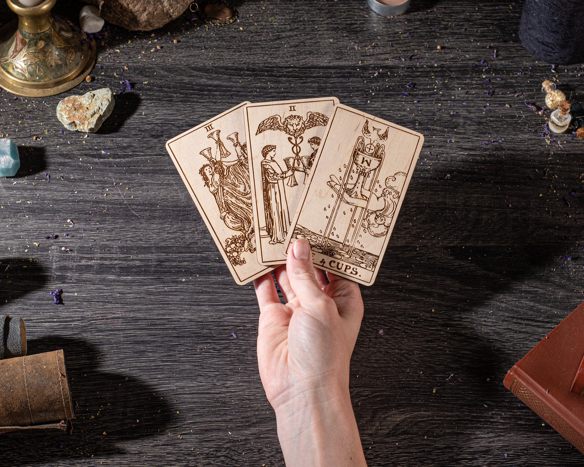 A hand is holding three wooden tarot cards against a dark wooden background, with various objects such as crystals, books, and a candle in the background.