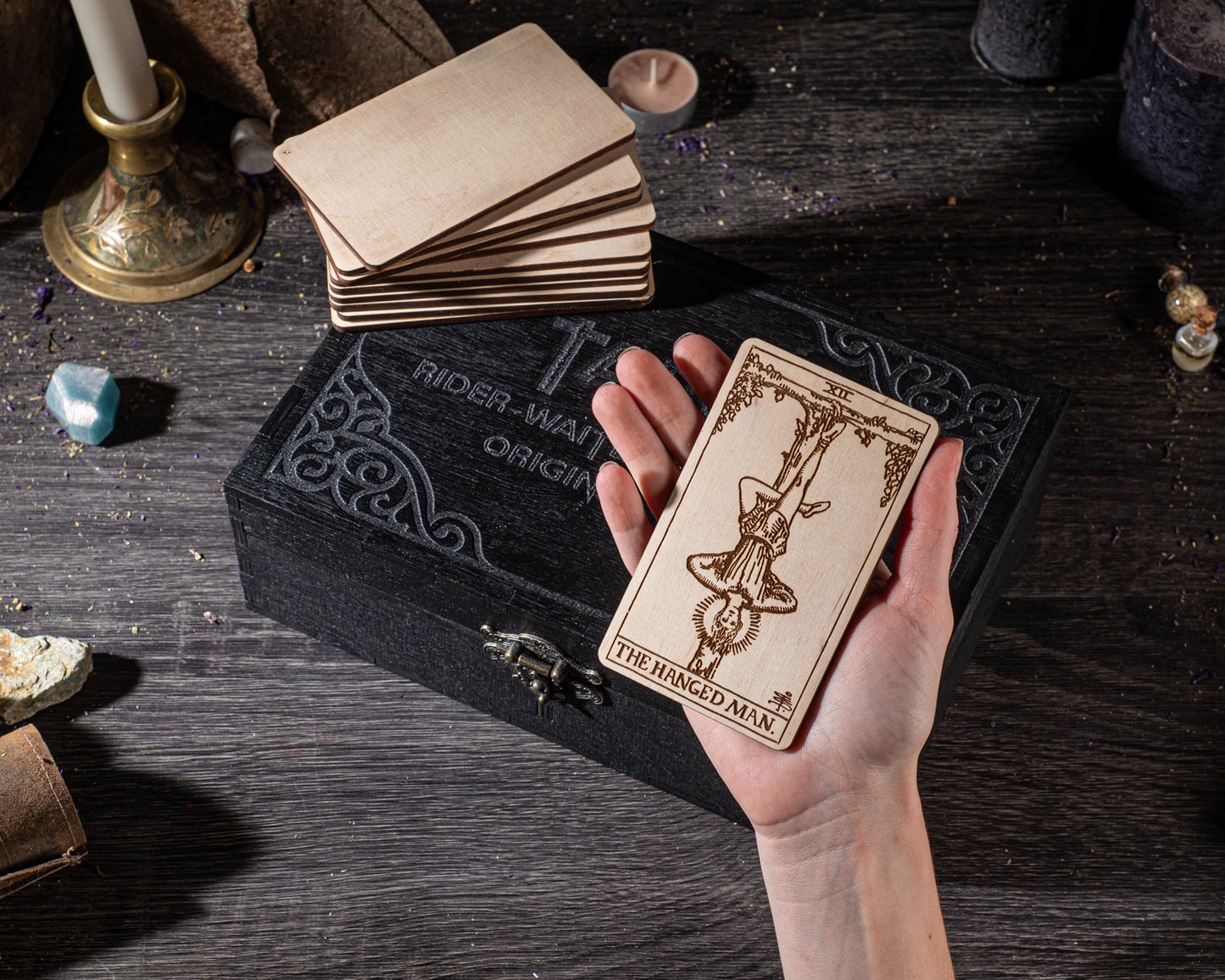 A hand holding a wooden tarot card with a design on it, placed on a black box with a decorative border. The box is surrounded by various objects, including candles, crystals, and other items, suggesting a mystical or spiritual setting.
