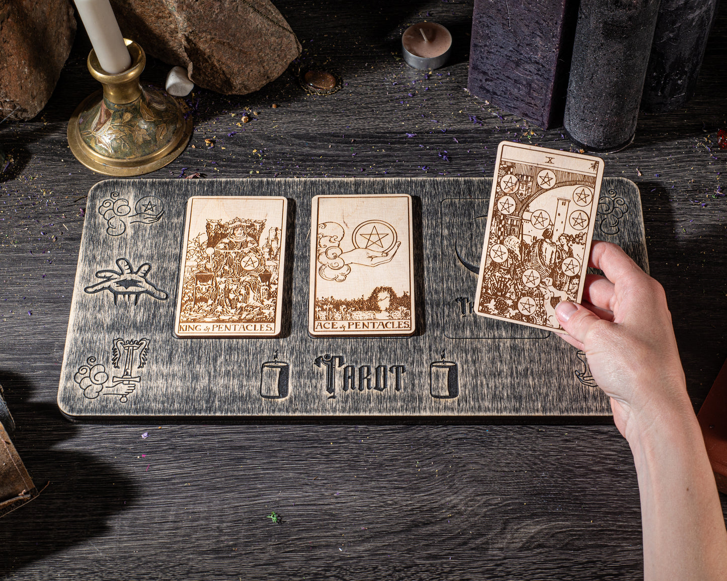A hand is holding a tarot card over a wooden board with three other tarot cards on it, placed on a dark wooden surface.
