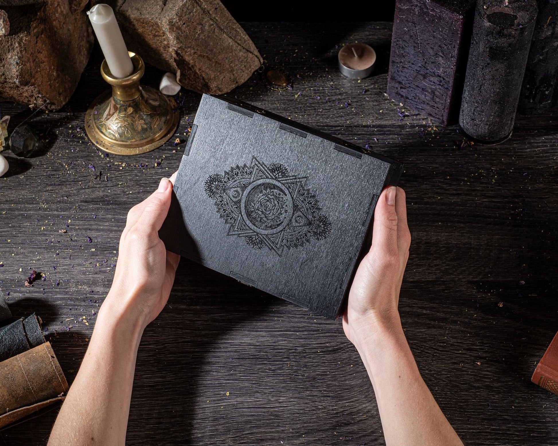 A person's hands holding a black book with an intricate design on the cover, placed on a wooden surface with various objects and candles in the background.