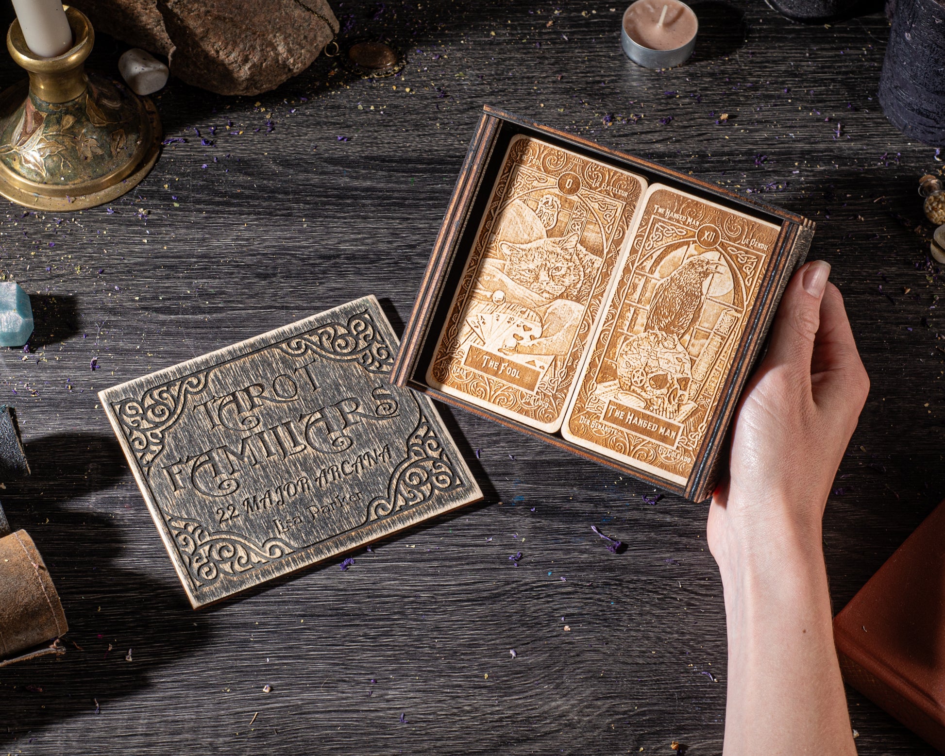 A hand is holding a wooden box containing two tarot cards, one open and one closed, on a dark wooden surface.