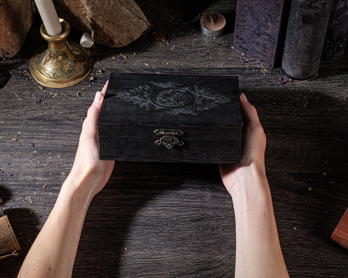 A pair of hands holding a black box with an intricate design on it, placed on a wooden surface with various objects and candles in the background.