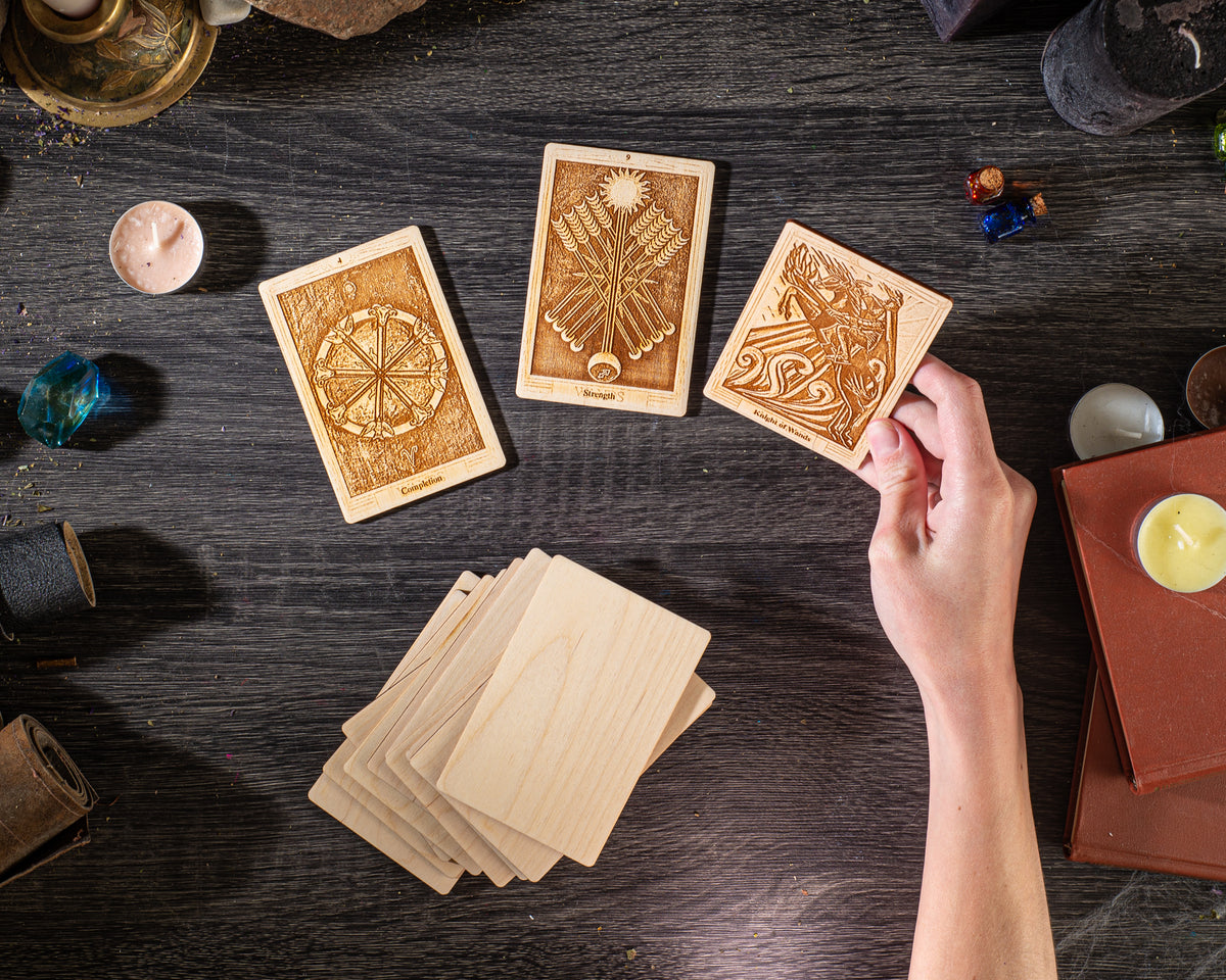 a person's hand holding a wooden card with an intricate design, surrounded by several other wooden cards and various objects on a dark wooden surface.