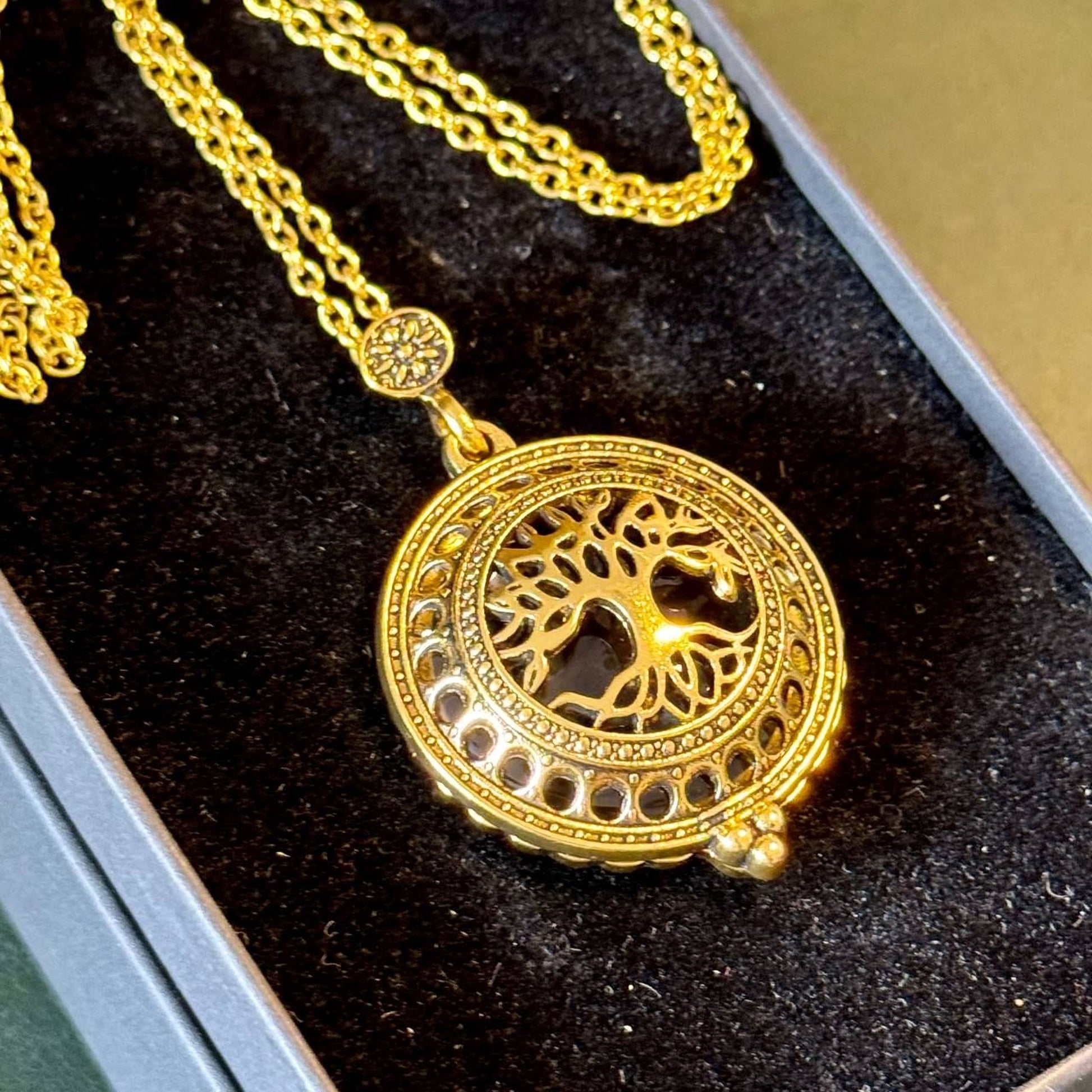 A gold-colored pendant necklace with a circular design featuring a tree motif, displayed in a black box.