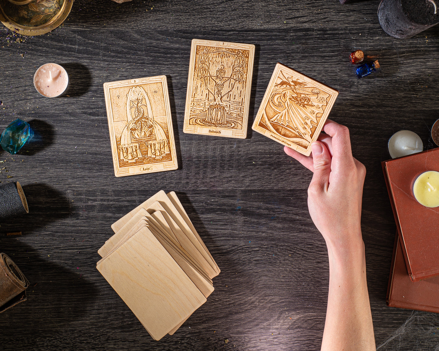 a hand holding a wooden card with an illustration on it, surrounded by various objects such as candles, books, and other items on a wooden surface.