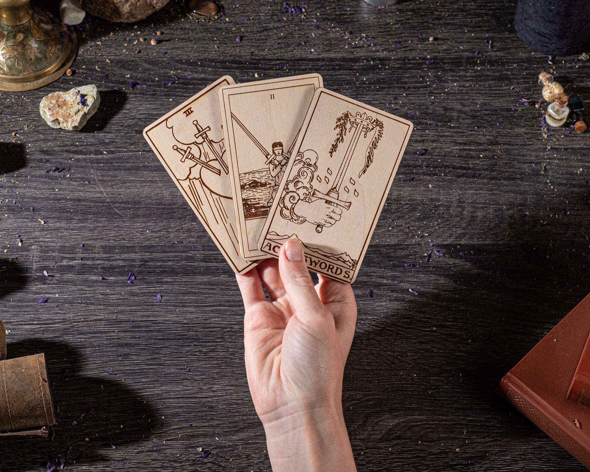 A hand is holding three wooden tarot cards against a dark background, with various objects scattered around.