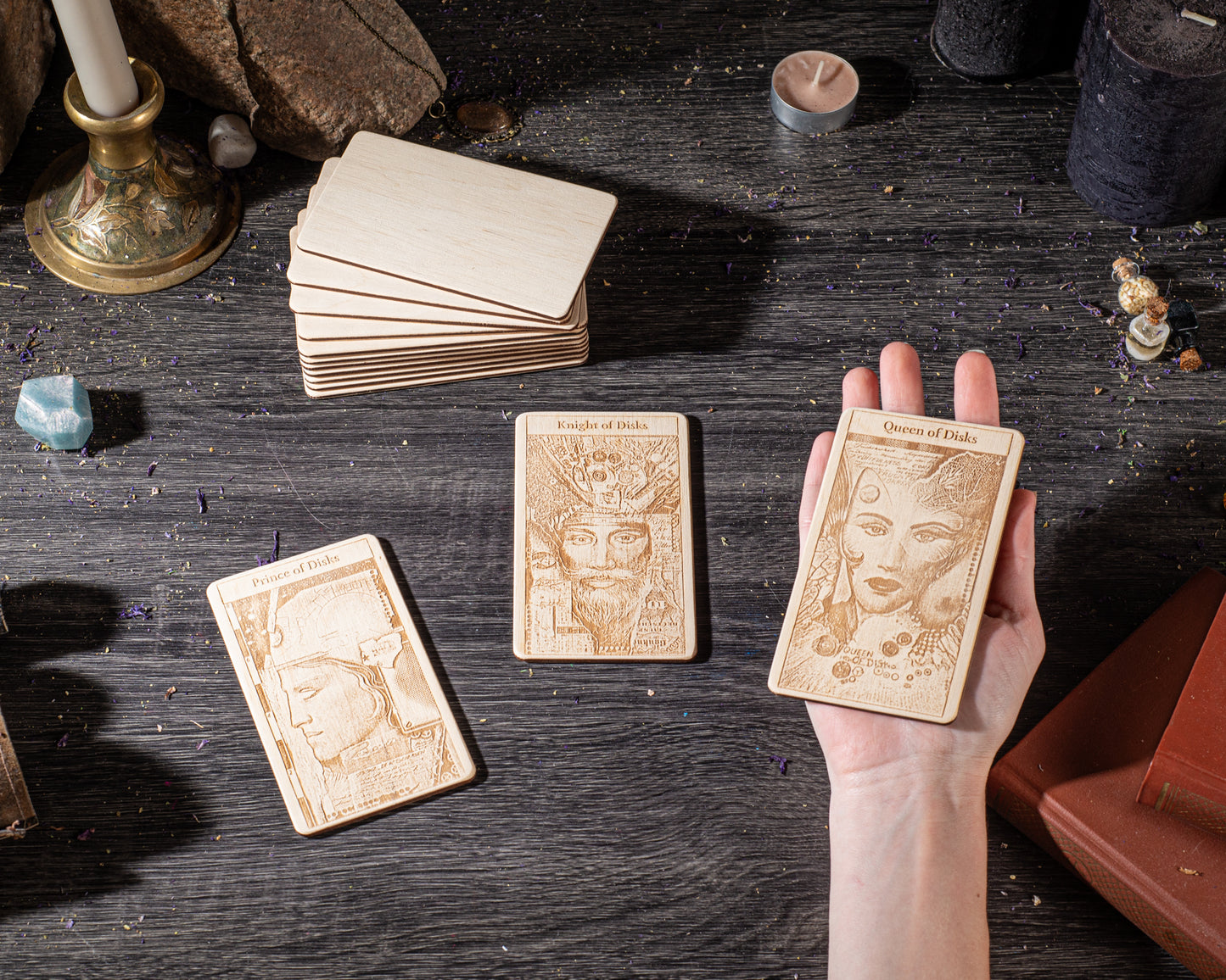 A hand is holding three wooden tarot cards on a dark wooden surface, surrounded by various objects such as candles, crystals, and books.